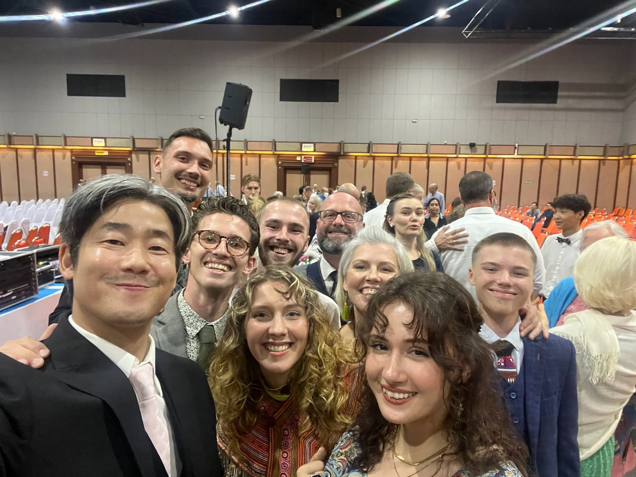 Group of people dressed in formal attire taking a selfie at a large event hall with orange seating.
