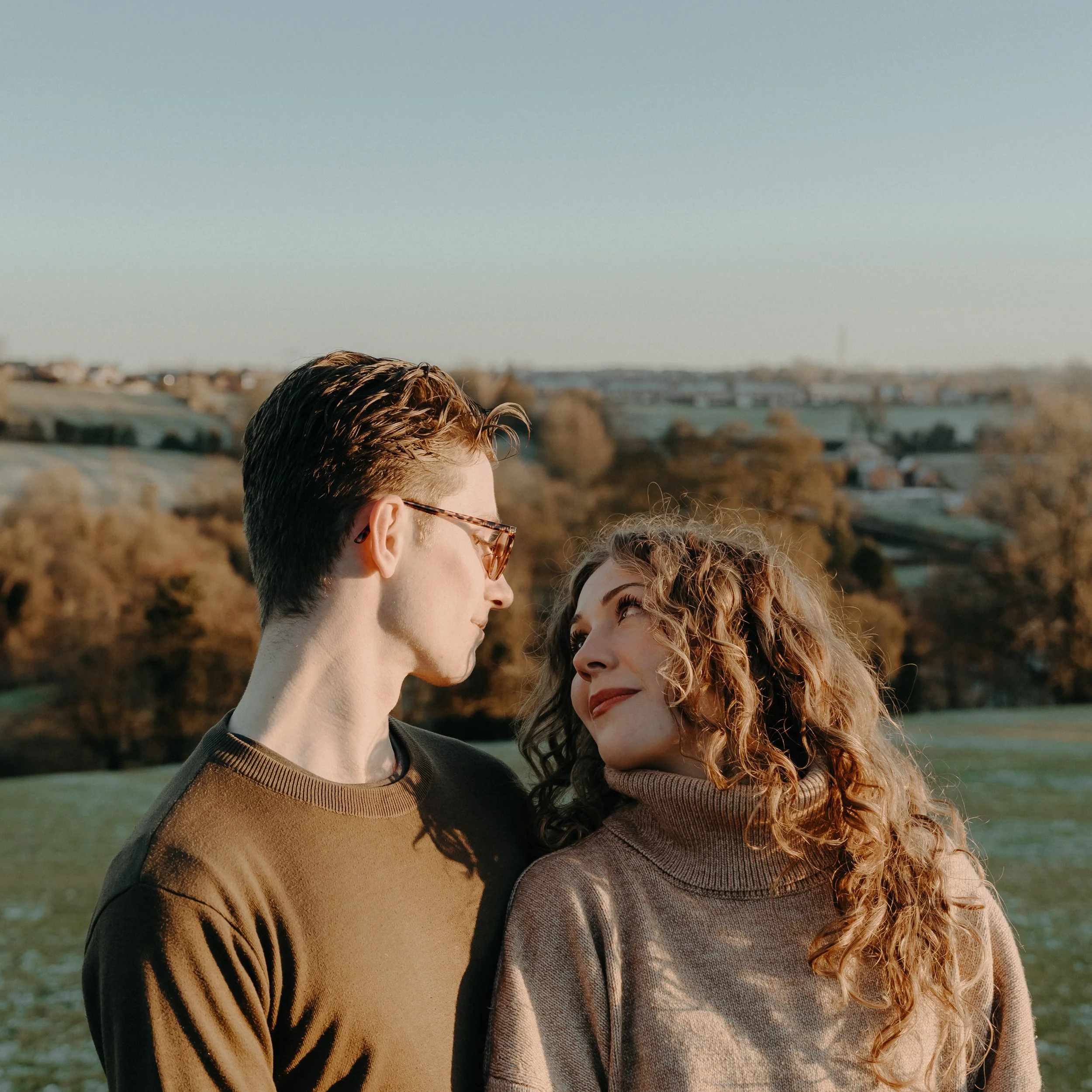 A young man and woman in an outdoor setting looking at each other. The man has short, dark hair and wears glasses and a brown sweater. The woman has curly, light-colored hair and wears a beige turtleneck sweater. They are standing on a grassy field w