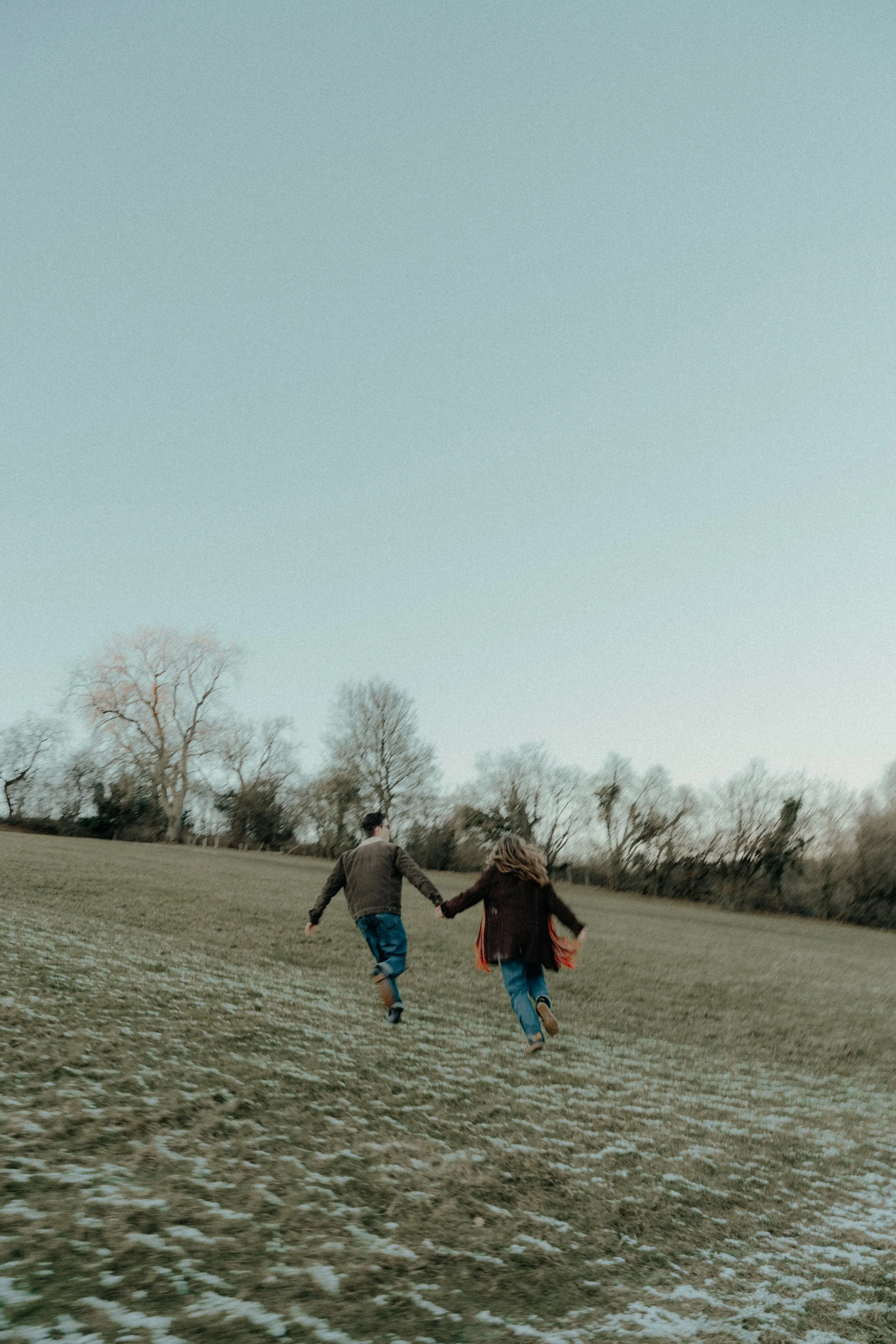 Two kids, a boy and a girl, holding hands and running across a grassy field with sparse trees in the background under a clear sky.