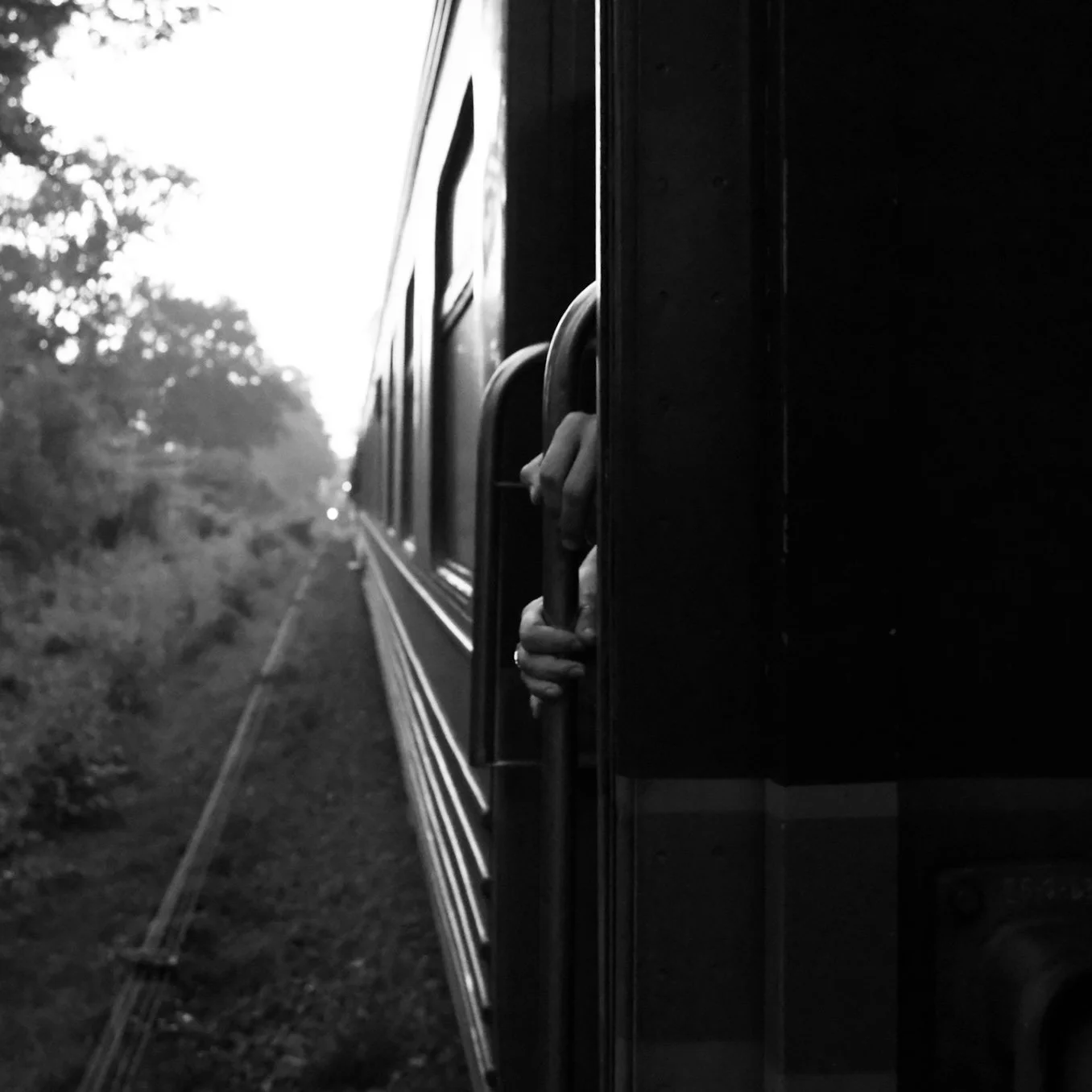 A black and white photo of a train moving along a track with trees in the background. Someone's hand is gripping a handle outside the train window.