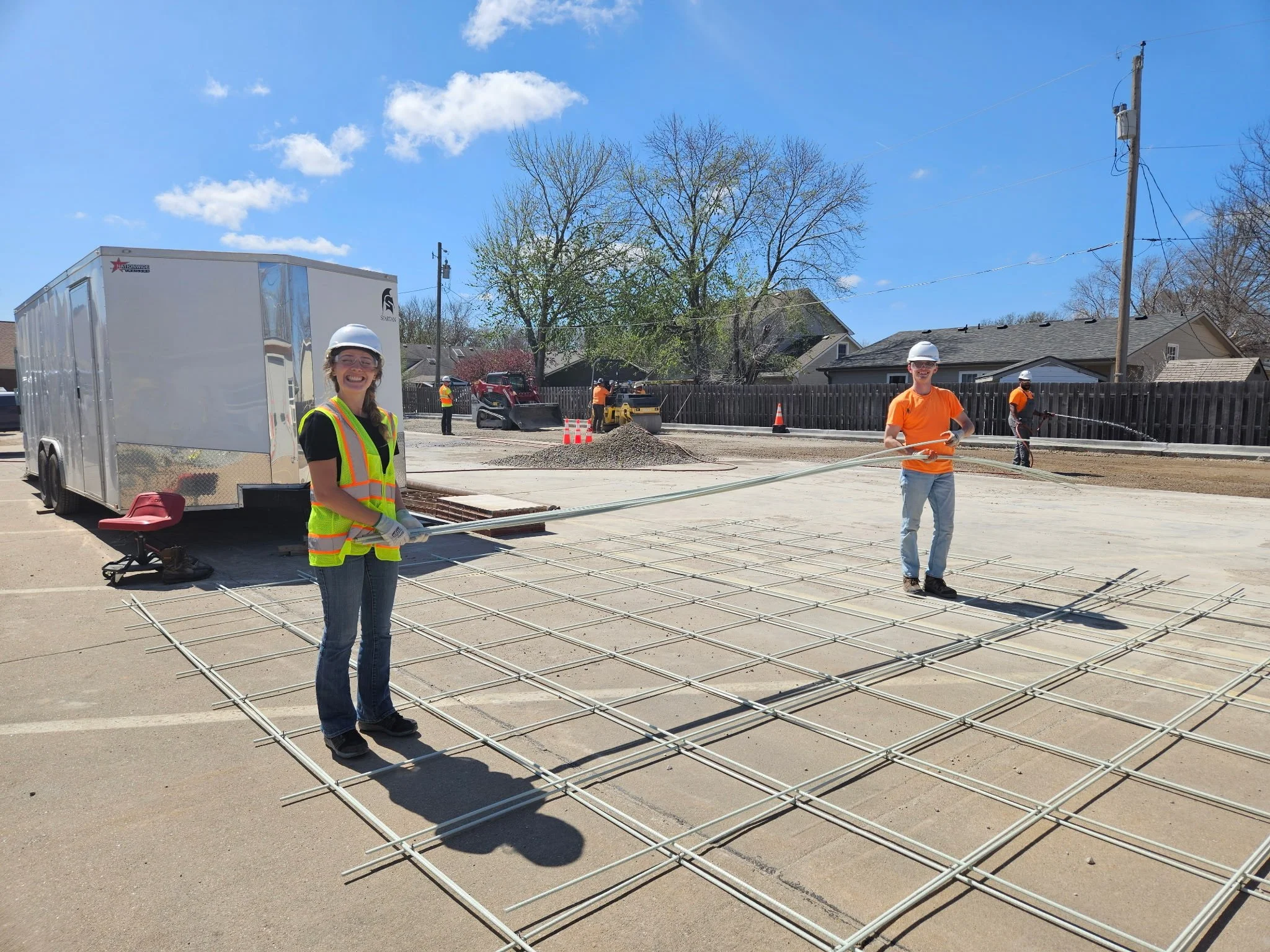 Construction workers setting foundation rebar on a parking lot with a bright blue sky and some trees in the background.