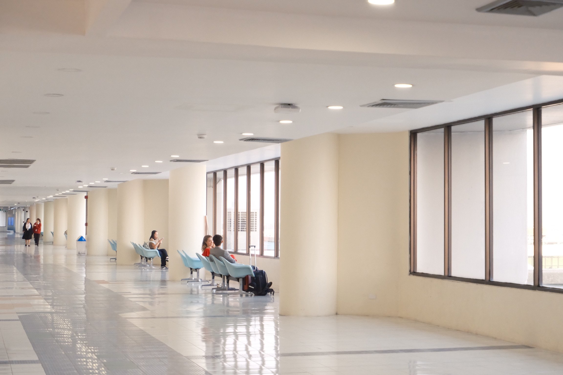 Empty waiting area in an airport with light blue chairs, large windows, and a few travelers sitting and using their phones.