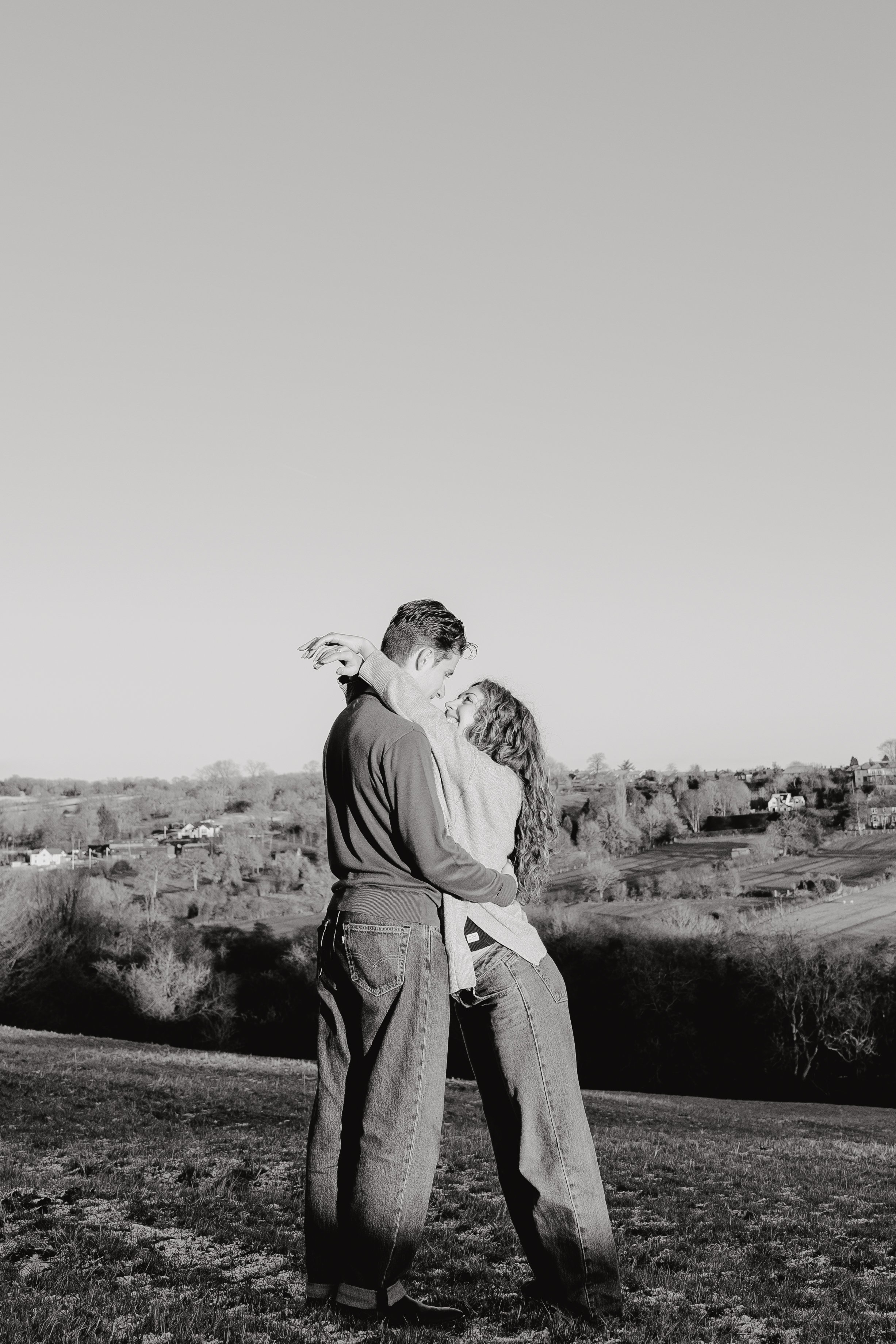 A black and white photo of a couple embracing outdoors, with a landscape of trees and houses in the background.