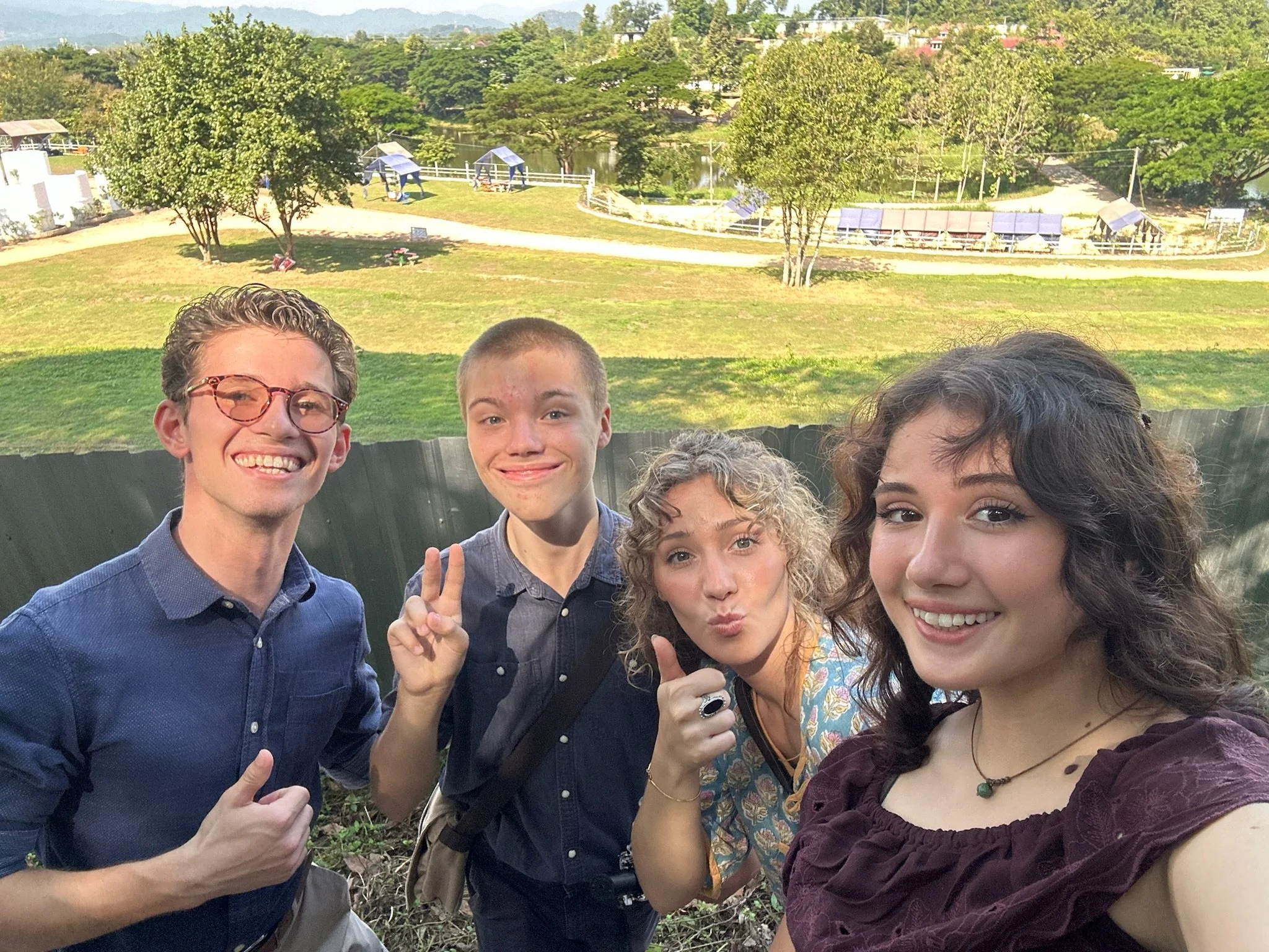 Four young people posing outdoors for a selfie in a park, with green grass, trees, and a pond in the background.
