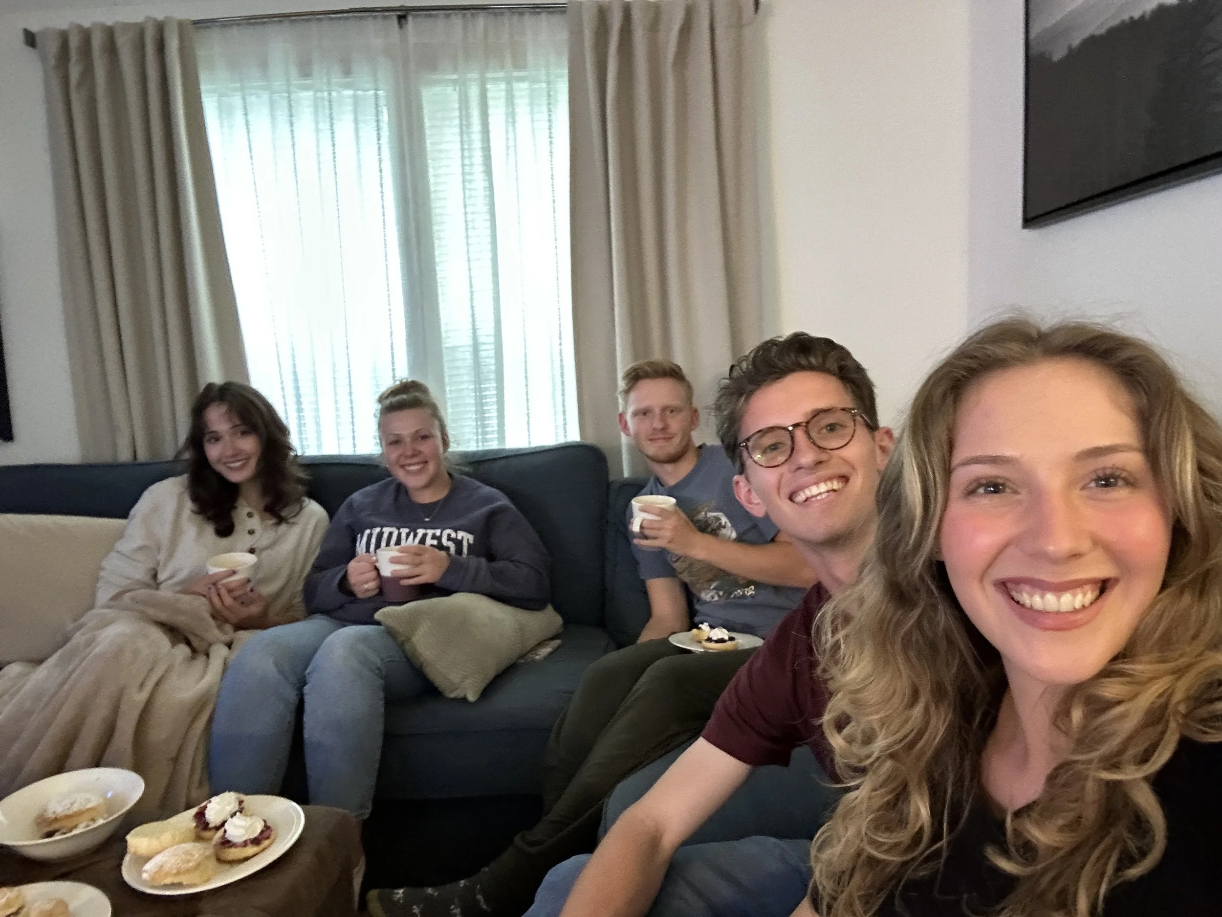 Group of five young adults sitting on a couch, smiling, with food and drinks, in a living room with curtains over windows.