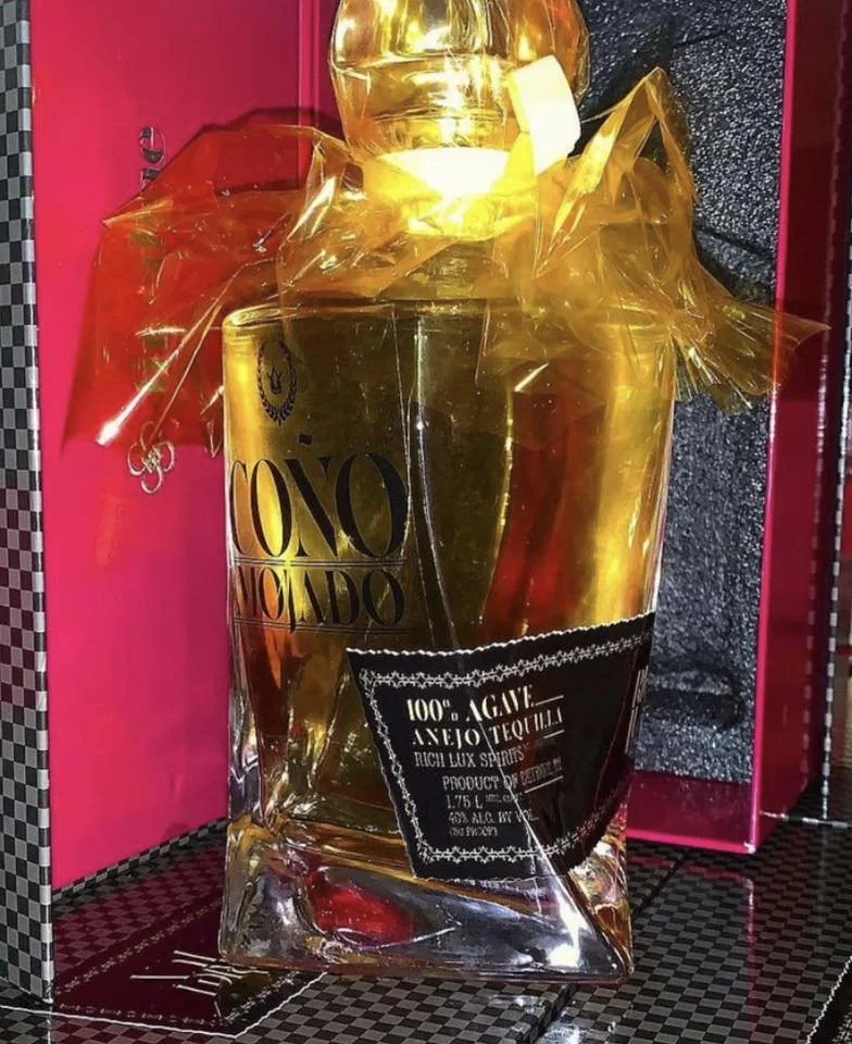 A large, gold-colored liquor bottle, wrapped in clear plastic with black and gold text labels, set against a pink background.