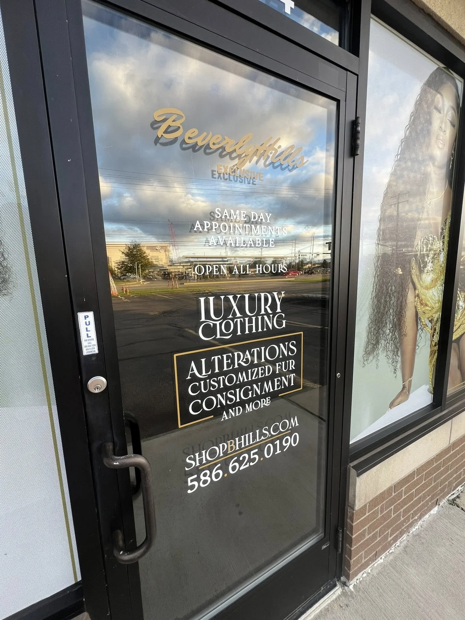 Glass storefront door with signage advertising Beverly Hills Exclusive luxury clothing, alterations, customized fur, and consignment, with a reflection of the parking lot and sky. A large poster of a woman with curly hair and gold accessories is besi