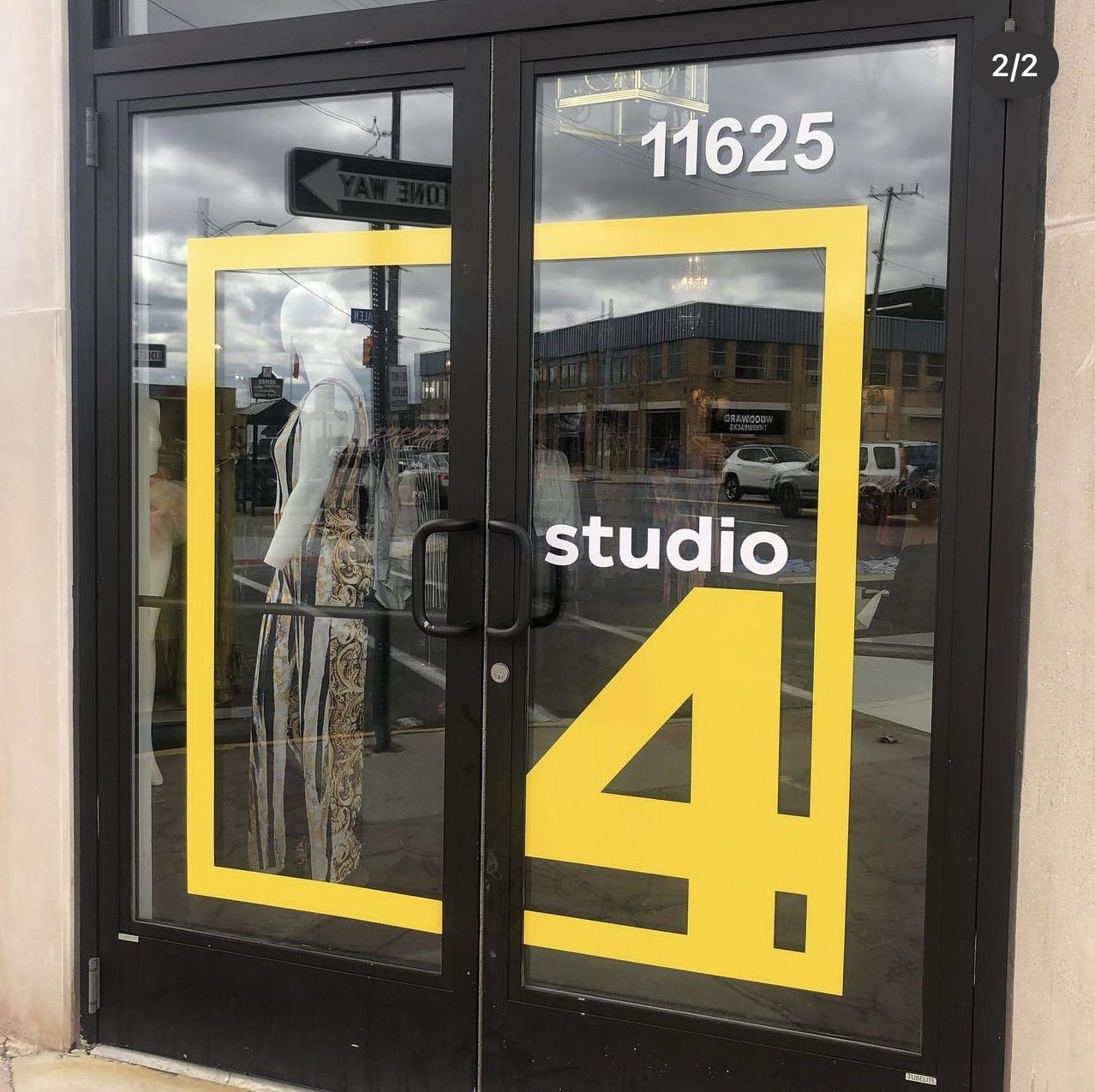 Glass door with black frame displaying the words 'studio 4' in white and a large yellow number 4. Reflection of street scene with parked cars and buildings outside.