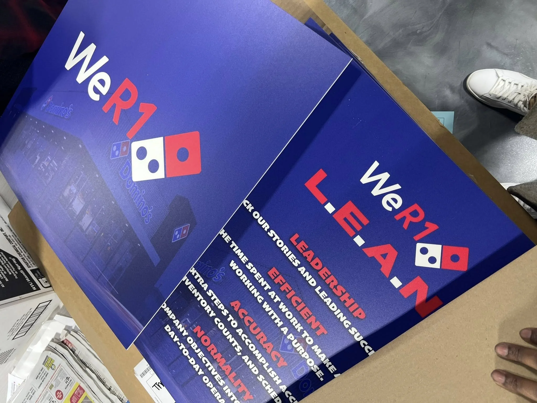 A large blue poster promoting the We R1 LEEA-N pizza franchise, featuring text about leadership, teamwork, and excellence, with Domino's logo and images of a pizza oven and pizza boxes.