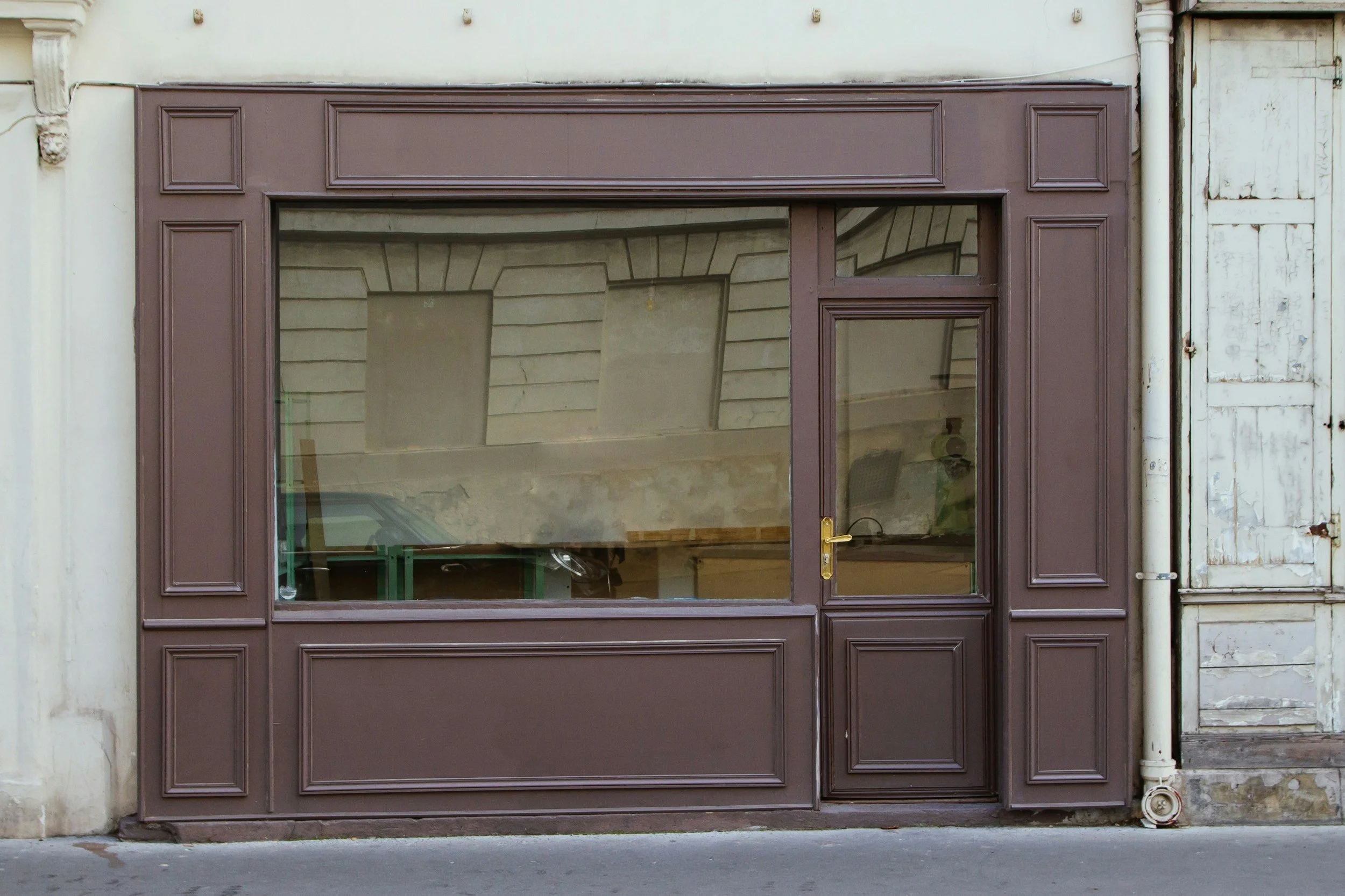 Brown storefront window with a small door, framed by decorative panels, on a city sidewalk.