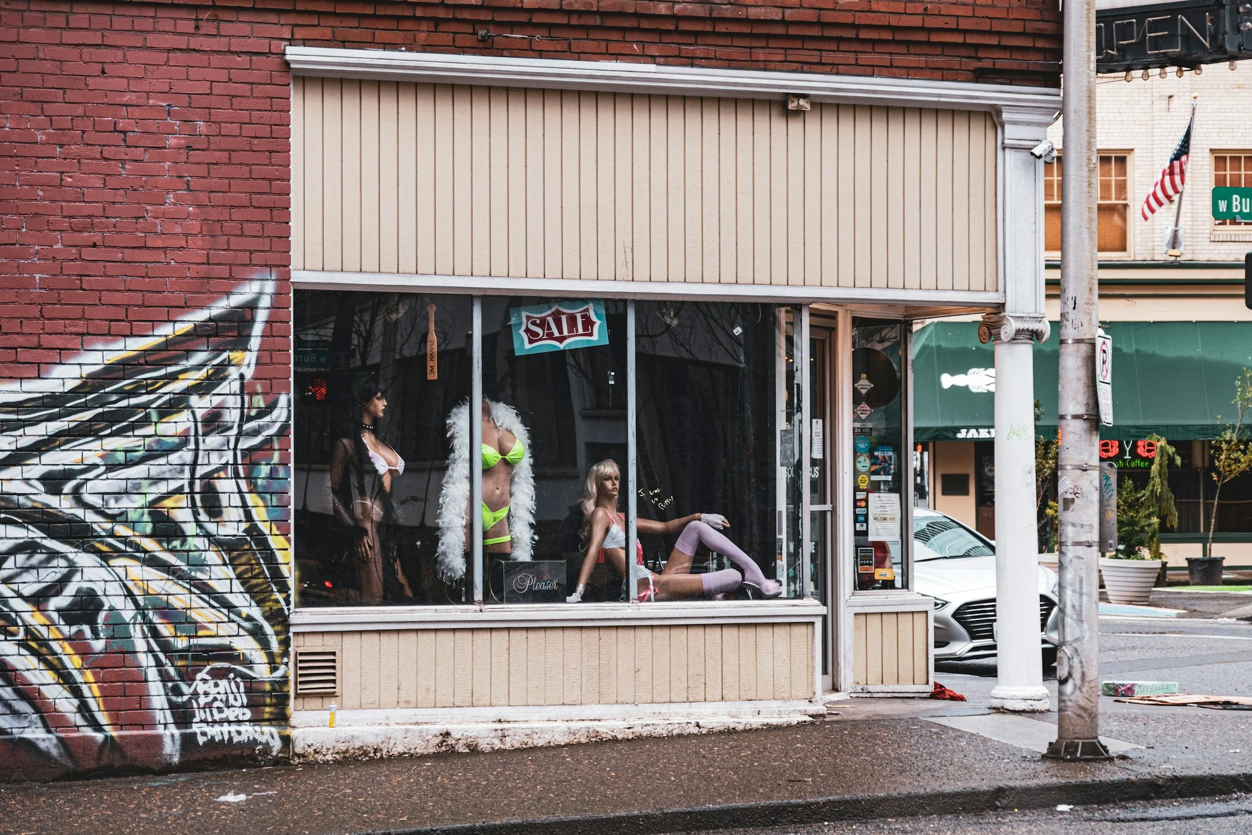 Storefront with mannequins in the window dressed in revealing clothing, including one in a neon bikini with a white feather boa, and a sale sign displayed in the window. There is colorful graffiti on the brick wall to the left.