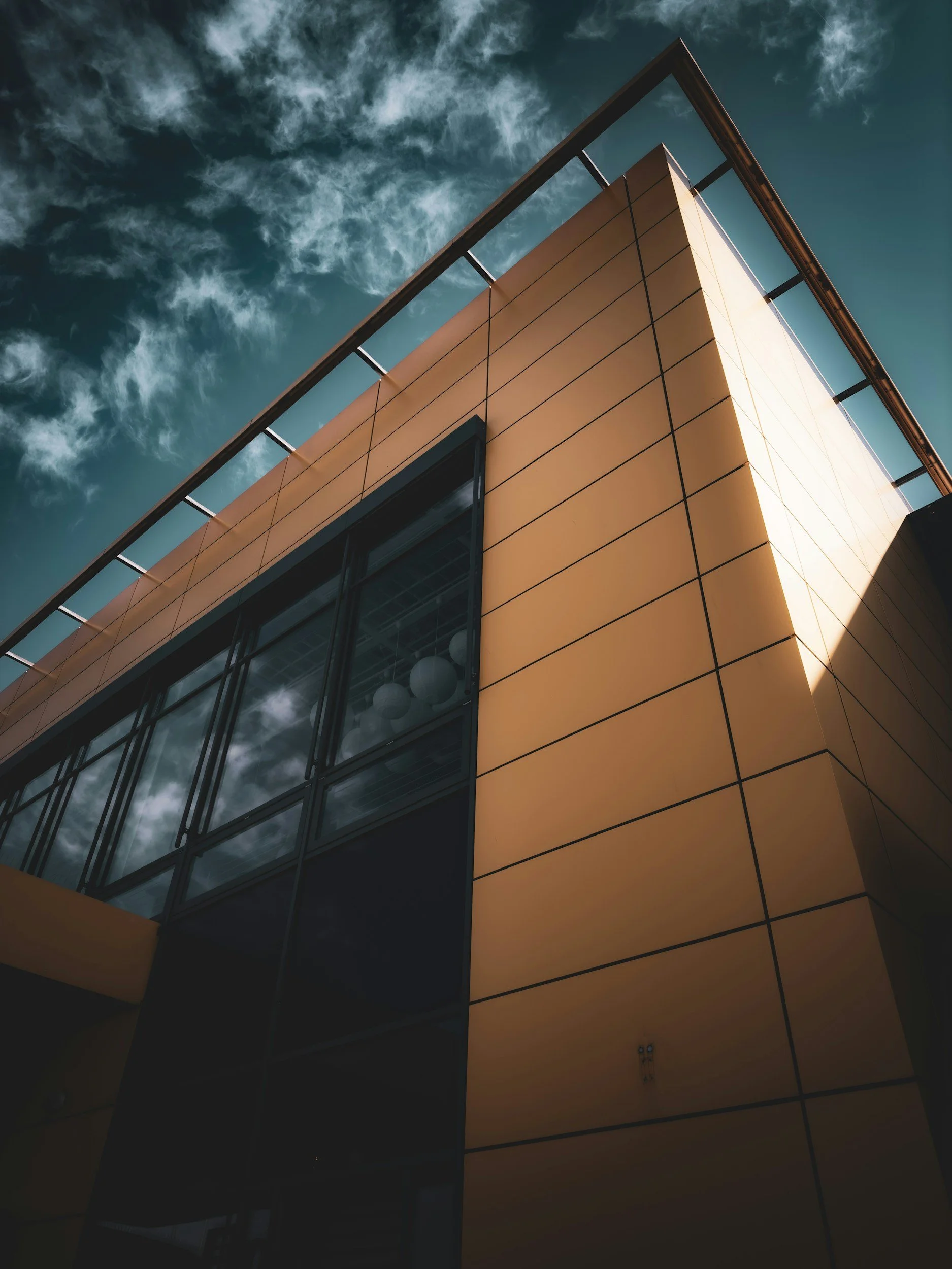 Low-angle view of a modern building with a yellow exterior, large glass windows, and a metal railing on the roof against a cloudy sky.