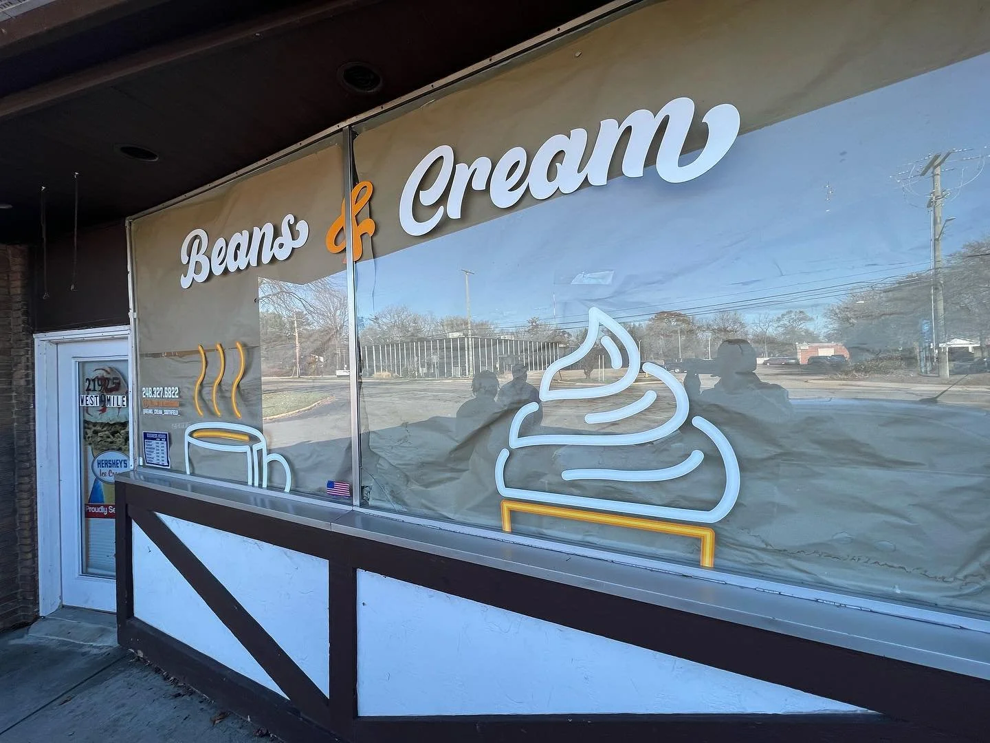 Storefront with neon sign reading 'Beans & Cream' featuring a steaming coffee cup and a swirl of cream, with a window reflecting the street scene outside.