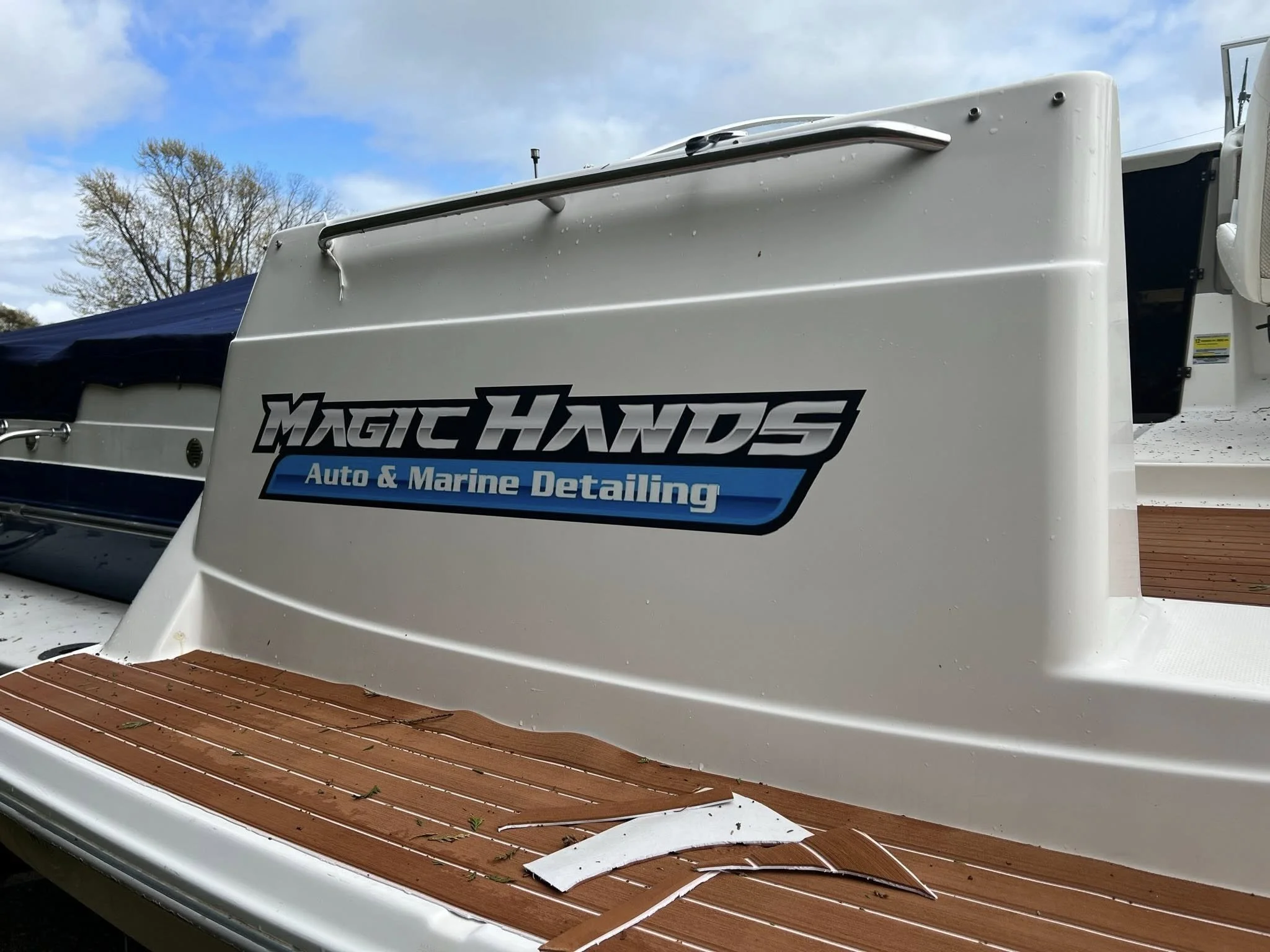 Close-up of a boat with a white hull and a wooden platform, with a logo that reads 'Magic Hands Auto & Marine Detailing' on the side.