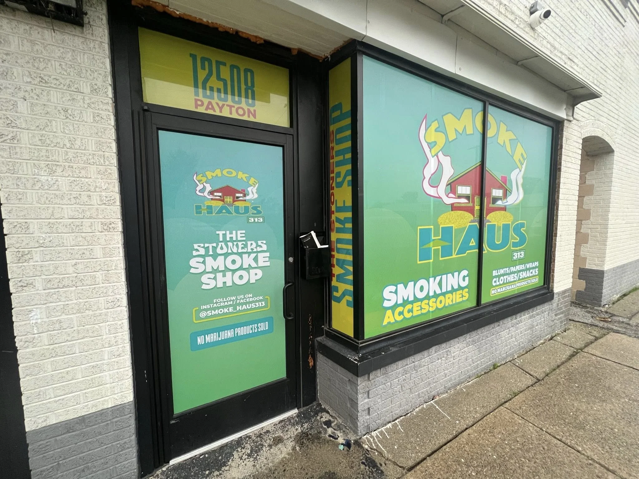 Storefront window and door advertising a smoke shop named Smoke Haus, featuring the store's name, logo of a house with smoke rising, and text promoting smoking accessories, blunts, papers, wraps, clothes, snacks, and no marijuana products sold, locat