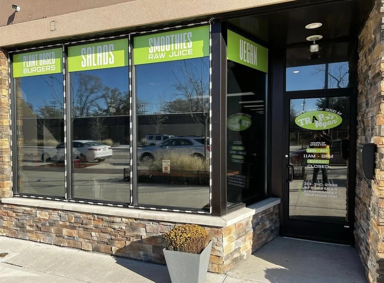 Storefront with large glass windows displaying signs for plant-based burgers, salads, smoothies, raw juice, and vegan options. The door has hours of operation, contact info, and vegan branding.