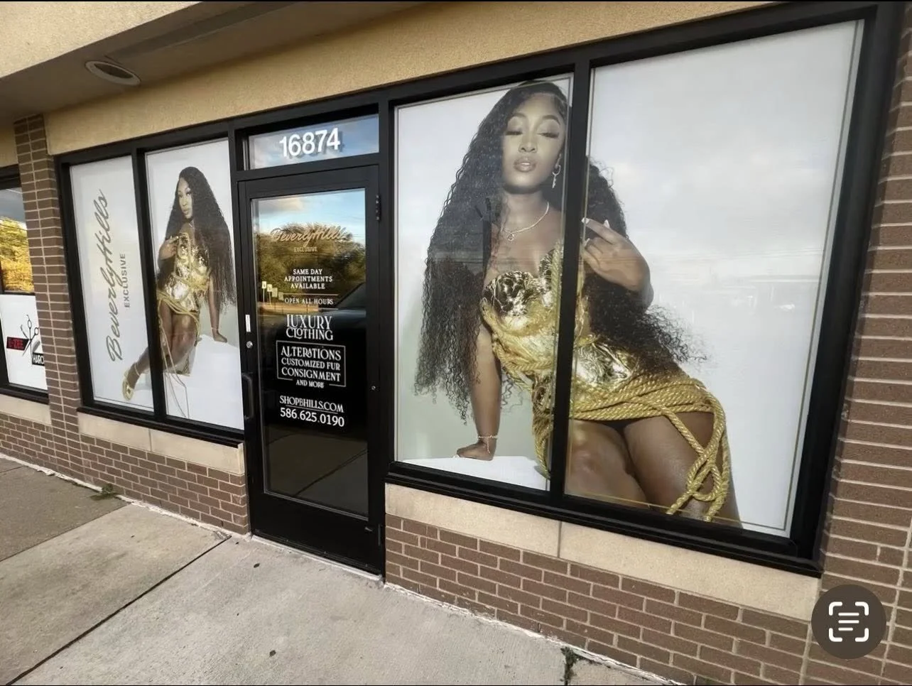 Storefront with large window graphics of a woman in a gold dress, advertising luxury clothing. Text on door: 'Same day appointments available, open all hours, luxury clothing, alterations, customized fur, consignment, shophillss.com, 586.625.0190.'