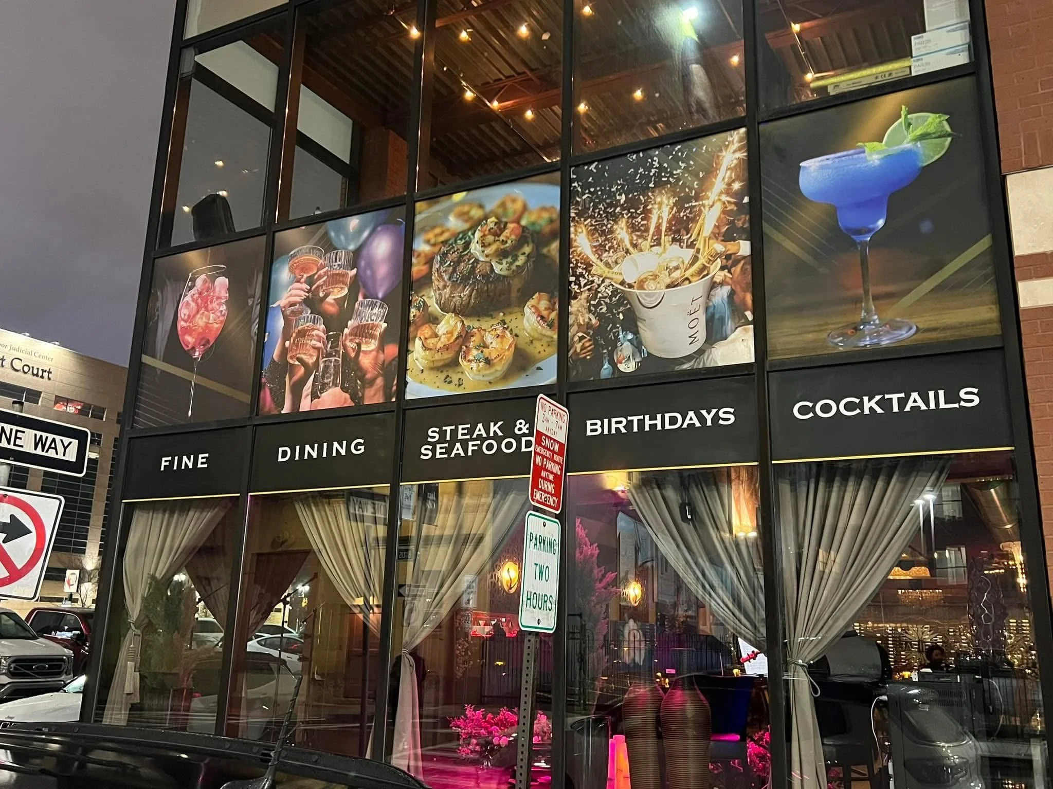 Exterior of a restaurant with large windows, displaying images of cocktails, food, and celebrations, with signs indicating fine dining, steak and seafood, birthdays, and cocktails.