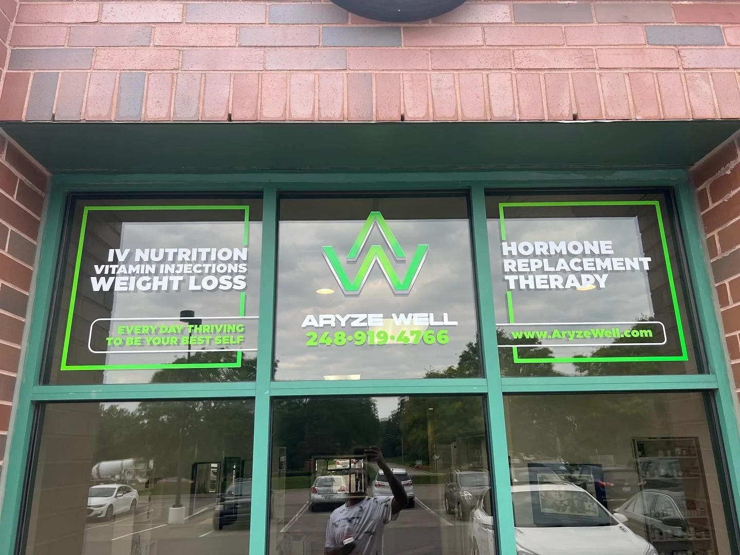 Storefront window with signs advertising IV nutrition, vitamin injections, weight loss, hormone replacement therapy, and website www.AryzeWell.com. The window features a green and white color scheme, with the name Aryze Well and phone number 248-919-