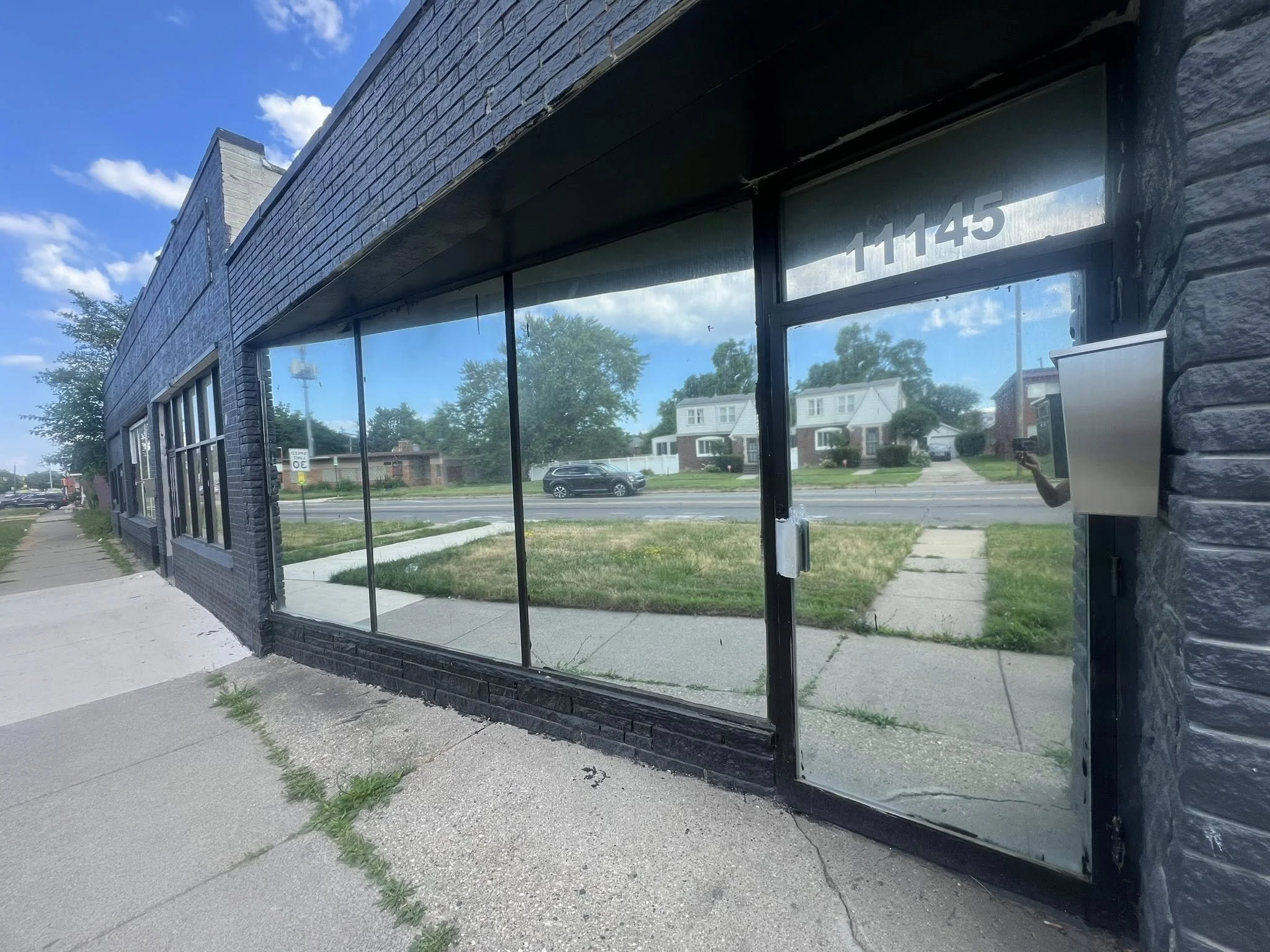 Storefront with black brick walls and large glass windows, street with cars and residential houses in the background, blue sky with some clouds.