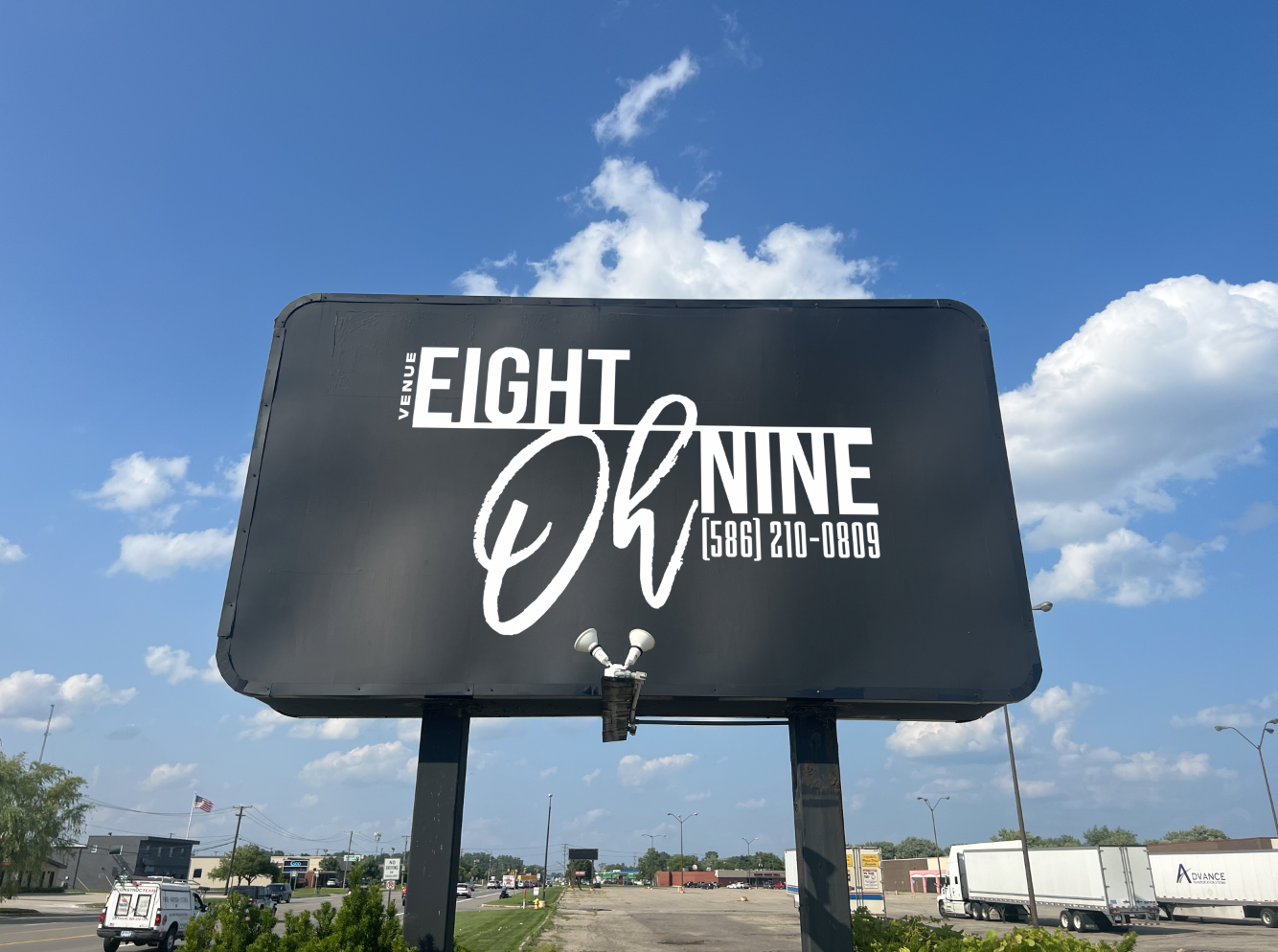 Large black billboard for EIGHT NINE Venue with contact number 586-210-0809, against a blue sky with scattered clouds, near a road with trucks and cars.