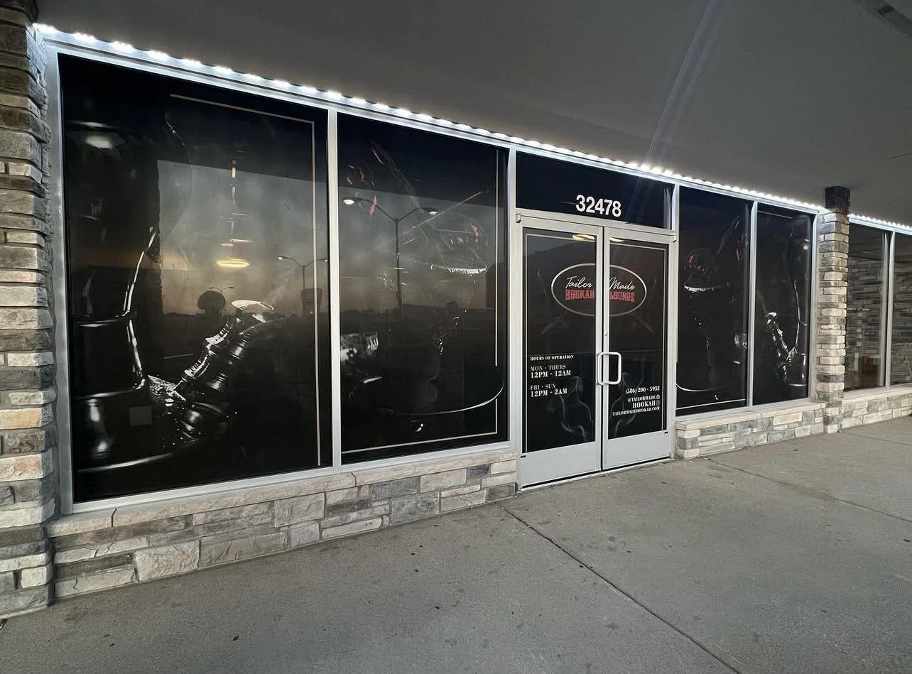 Front view of a storefront with large windows displaying images of motorcycle parts and a sign that reads "Tailored Made Hookah & Burger". The storefront has a stone facade and a black door with the store's hours.