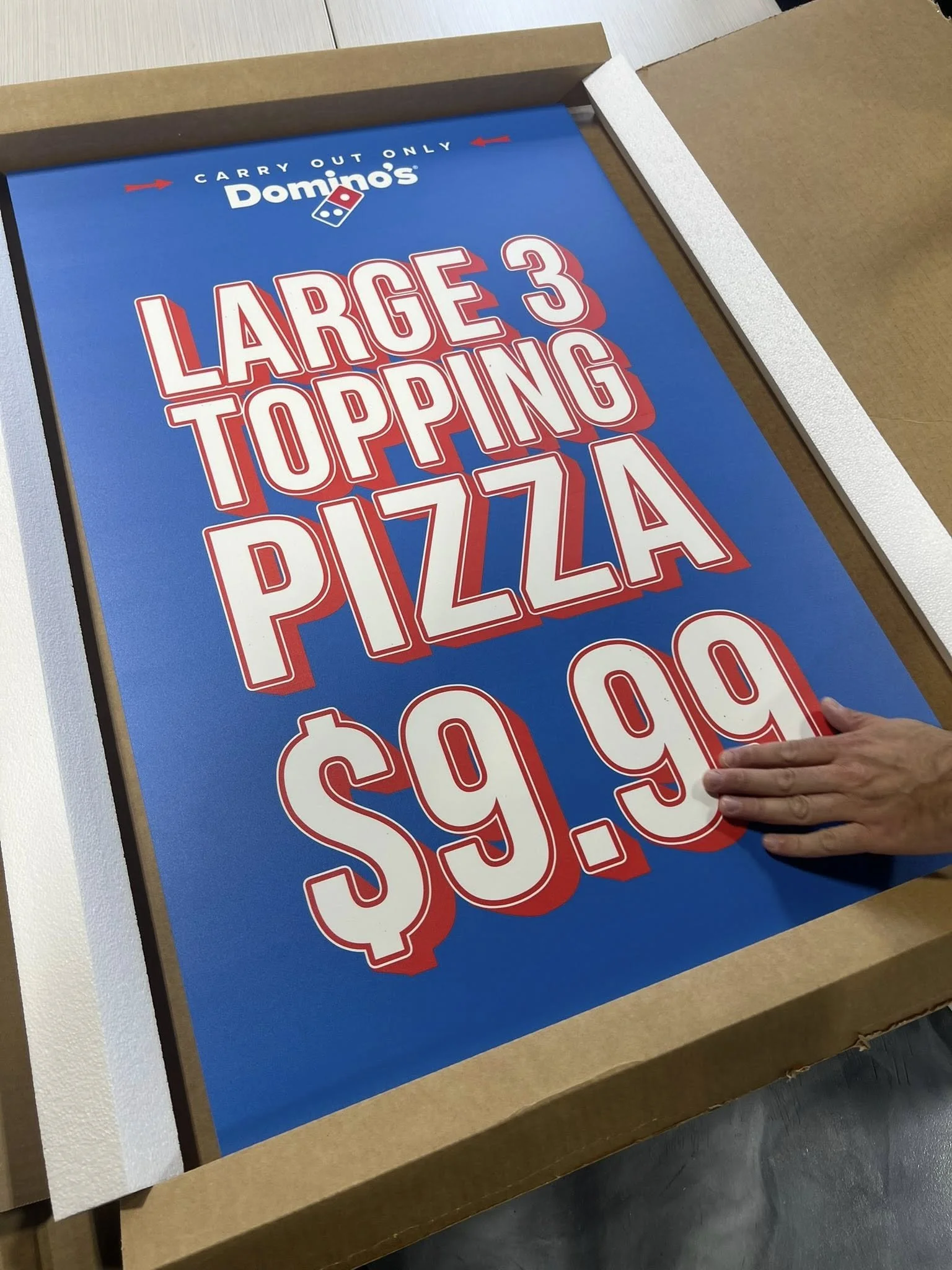 A blue sign advertising a large three-topping pizza for $9.99 at Domino's, with a person's hand touching the bottom right corner of the sign.
