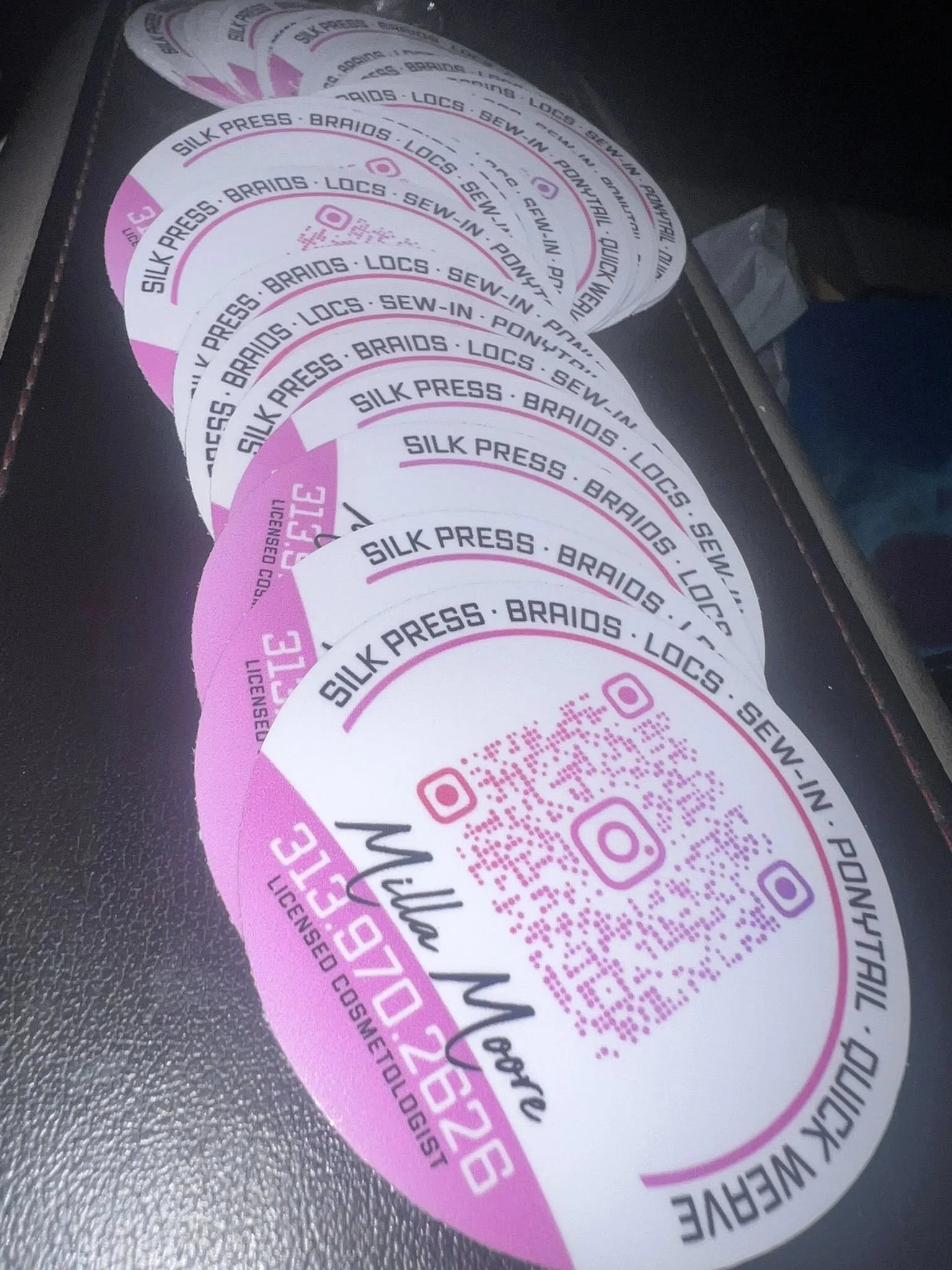 Multiple circular stickers with a pink and white design, featuring a QR code, Instagram logo, and text advertising Silk Press, Braids, Locs, Sew-In, Ponytail, and Licensed Cosmetologist, with an Instagram handle @MiraMoe and contact number 970-826-78