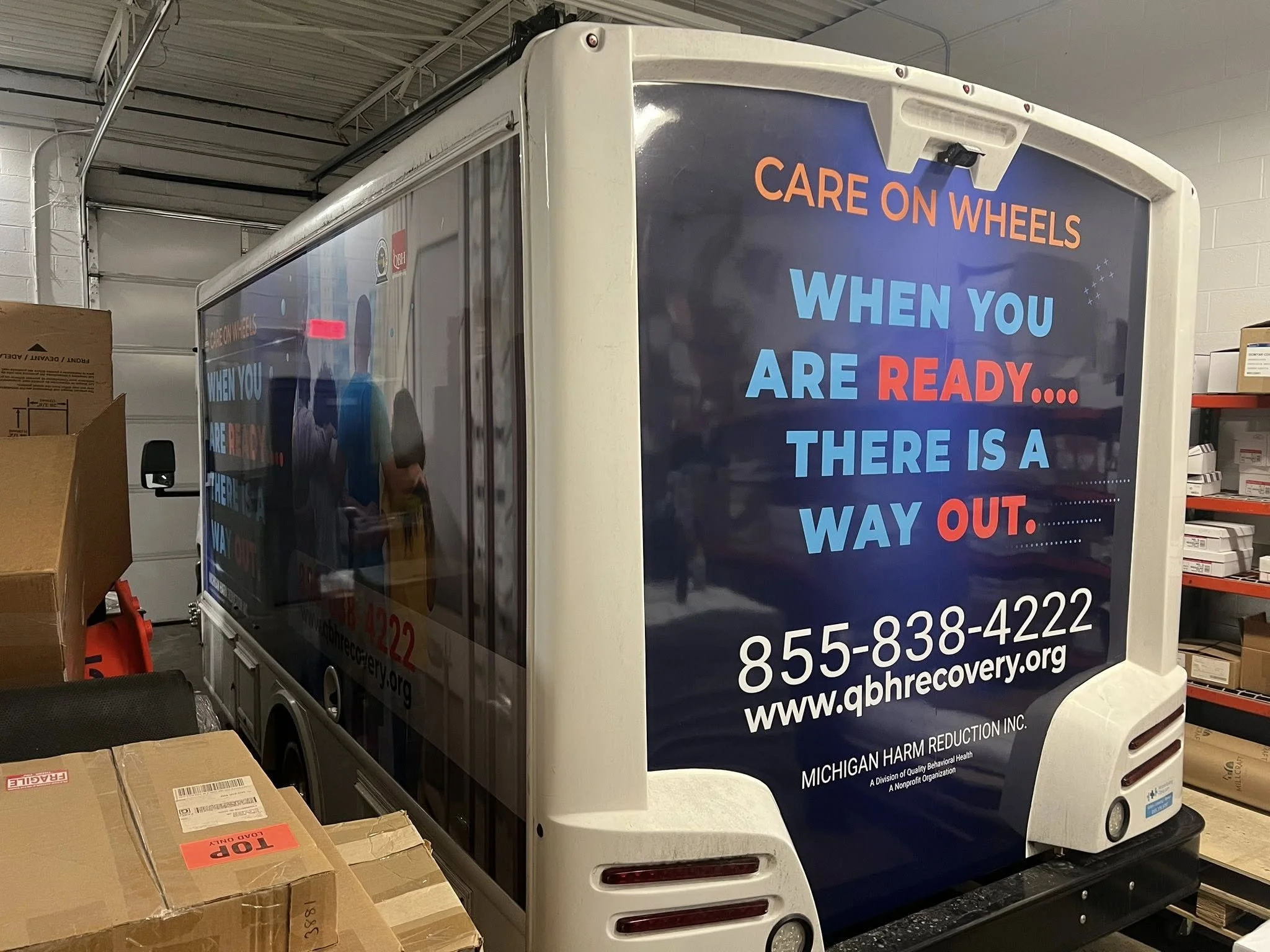 A mobile recovery service vehicle with an advertisement for a recovery organization that offers help when you're ready, providing a phone number and website, located inside a warehouse with cardboard boxes around.
