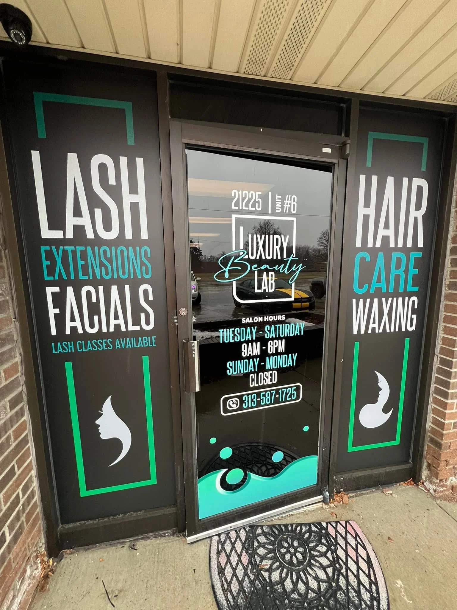 Front door of a beauty salon with signs advertising lash extensions, facials, hair care, and waxing services. Salon hours are Tuesday to Saturday from 9 AM to 6 PM, closed Sunday and Monday, with a phone number displayed.