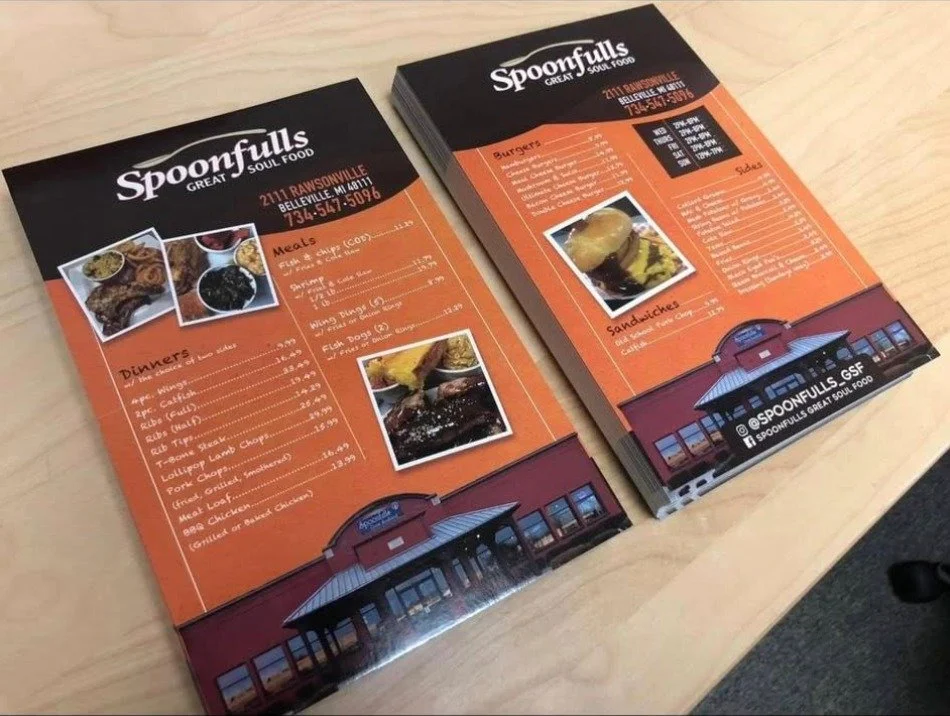 Two restaurant menus for Spoonfulls in Ransonville, displaying food and drink options with pictures of dishes on an orange background, placed on a wooden table.