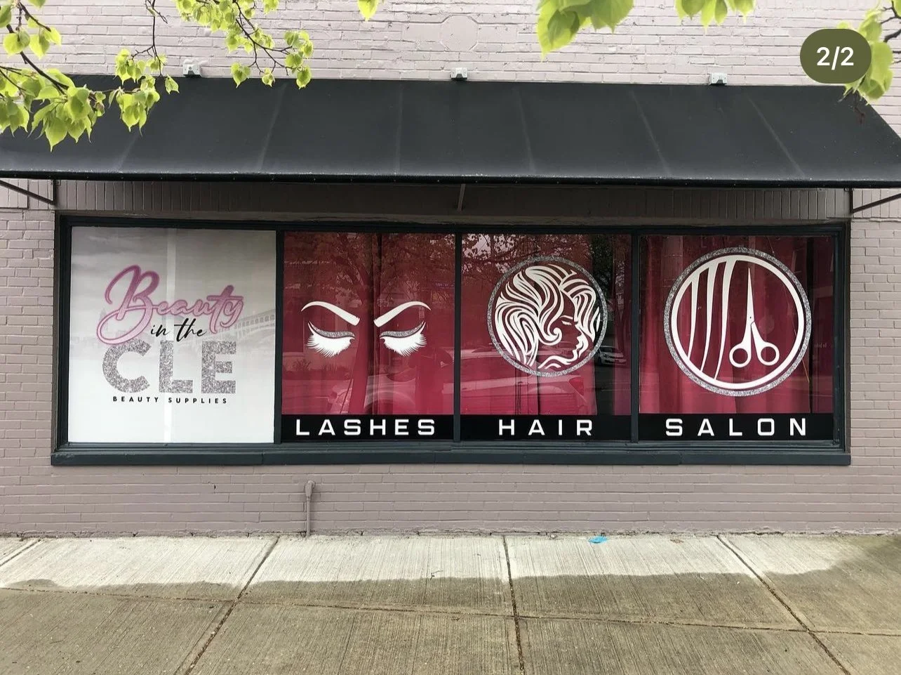 Storefront window advertising a beauty salon with images of eyes with long lashes, a woman's profile with styled hair, and barber scissors, with text reading "Beauty in the CLE, Lash, Hair, Salon, Beauty Supplies."