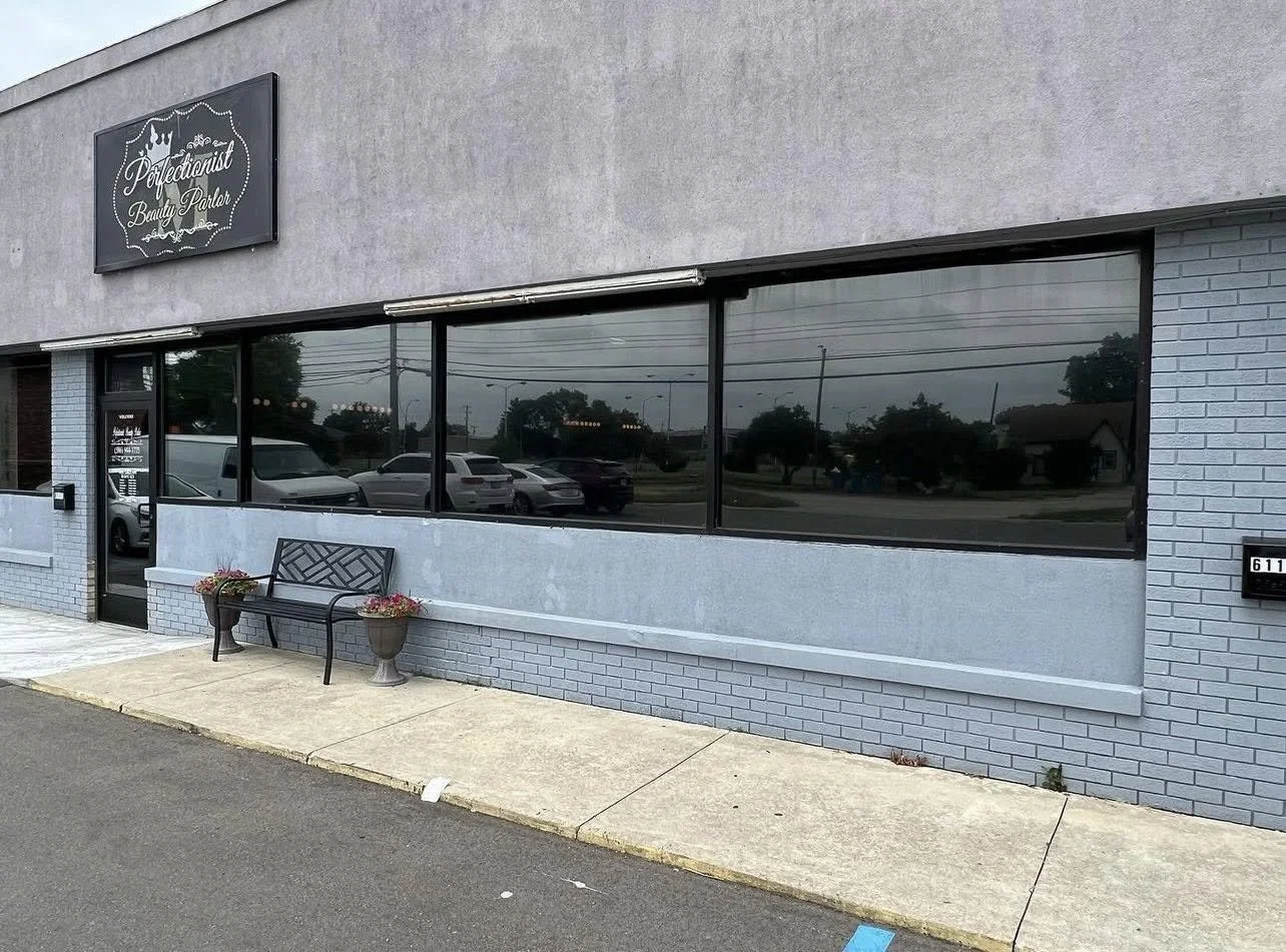 Exterior view of a beauty salon called 'Perfectionist Beauty Parlor' with large tinted windows, a bench with two flower pots outside, and a sidewalk in front.