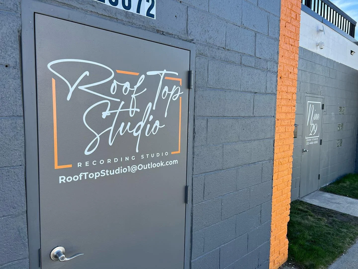 Outdoor view of a building with signs indicating Roof Top Studio Recording Studio and Nine 29 Event Studio. The main sign is on a gray door and includes an email address. The building has a dark gray painted brick wall with a vertical orange brick st