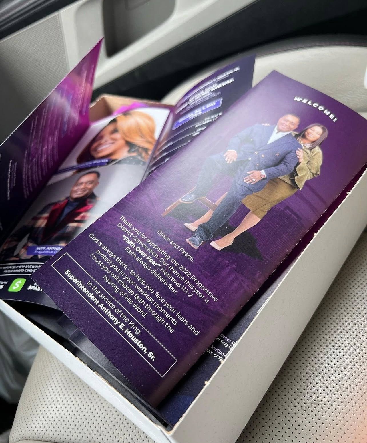 A pamphlet or program with a photo of a man and a woman sitting together, with text including a welcome message and a quote about faith and overcoming fear, placed on an airplane seat.