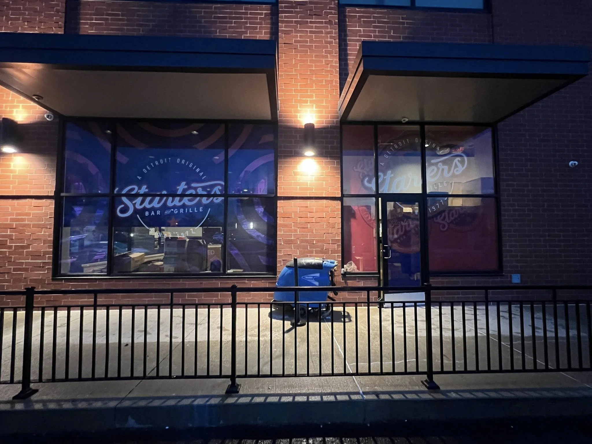 The exterior of a restaurant or bar named Stanley's, with brick walls, large glass windows, a lit sign, a door, a sidewalk, and a blue trash bin in front.