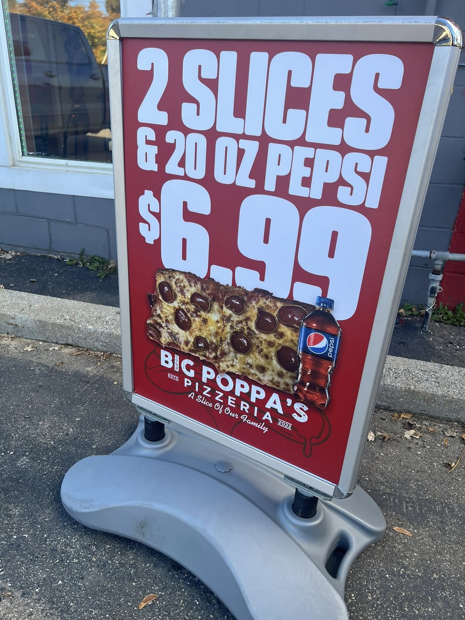 Outdoor sidewalk sign showing promotion for two slices of pizza and a 20 oz Pepsi for $6.99 at Big Poppa's Pizzeria, with an image of pizza and a Pepsi bottle included in the advertisement.