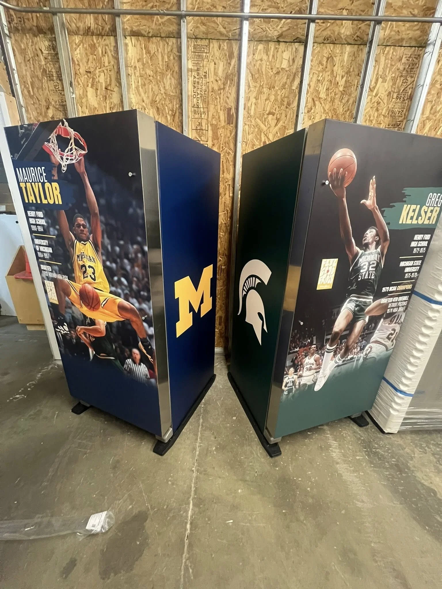 Two refrigerators with basketball player images and team logos, one for Michigan Wolverines and the other for Michigan State Spartans, placed side by side in an unfinished room.
