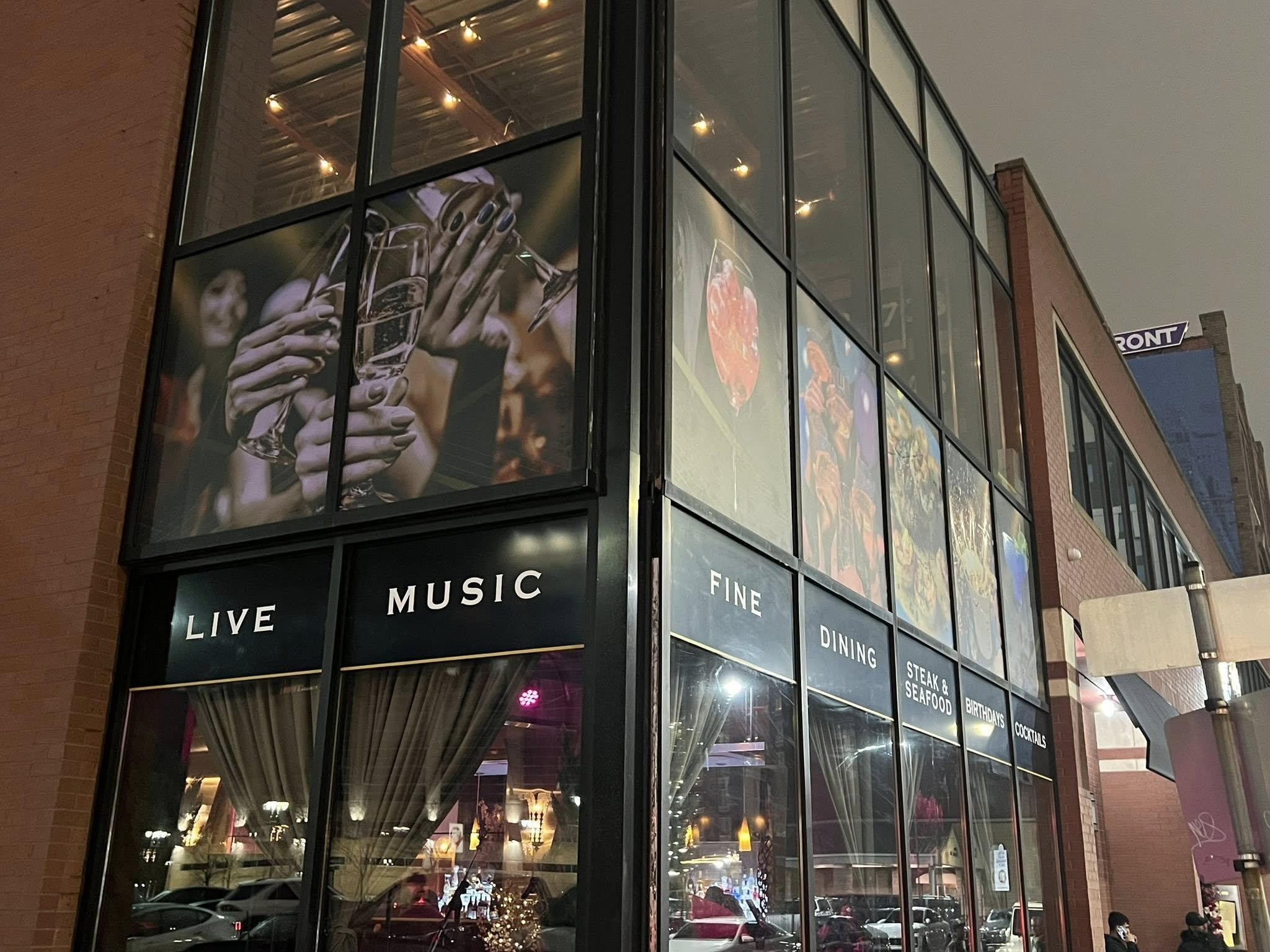 A storefront with large glass windows showing signs for live music, fine dining, steak and seafood, birthdays, and cocktails. The upper part of the building displays promotional images, including people clinking champagne glasses and images of food l