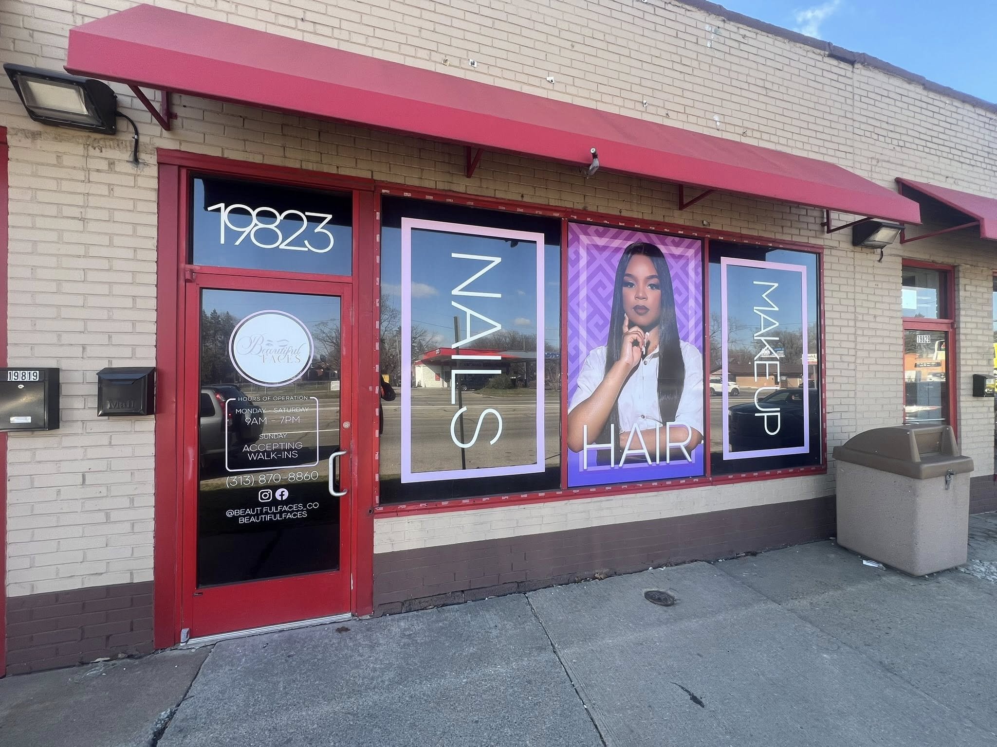 Exterior view of a beauty salon with red trim and awning. Large store window with promotional posters advertising hair styling services, including a woman with long, dark hair and dark lipstick. The door has the salon's name, operating hours, contact