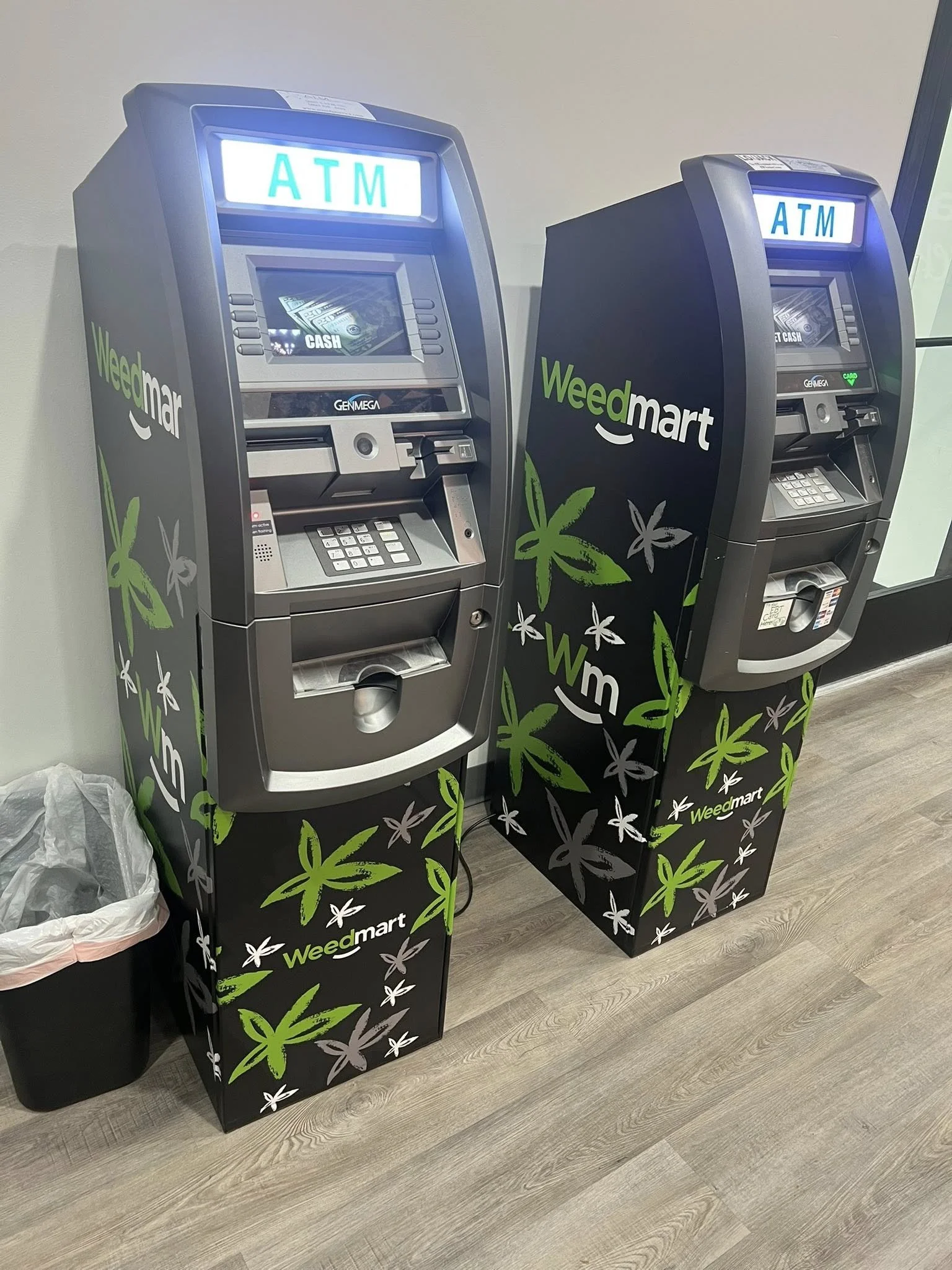 Two Weedmart ATM machines with green and white marijuana leaf designs on the black stands, located indoors on a wooden floor beside a trash can.