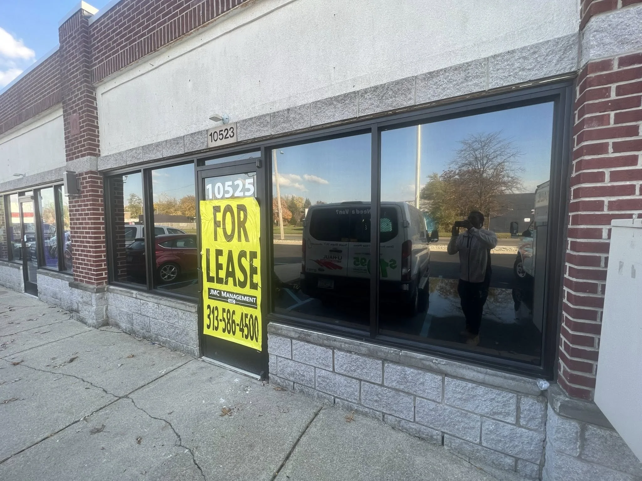 A commercial storefront with large glass windows and brick and stone exterior. There are two addresses visible, 10523 and 10525. A bright yellow sign in the window says "For Lease" with a phone number, 313-586-4500, and details about leasing manageme