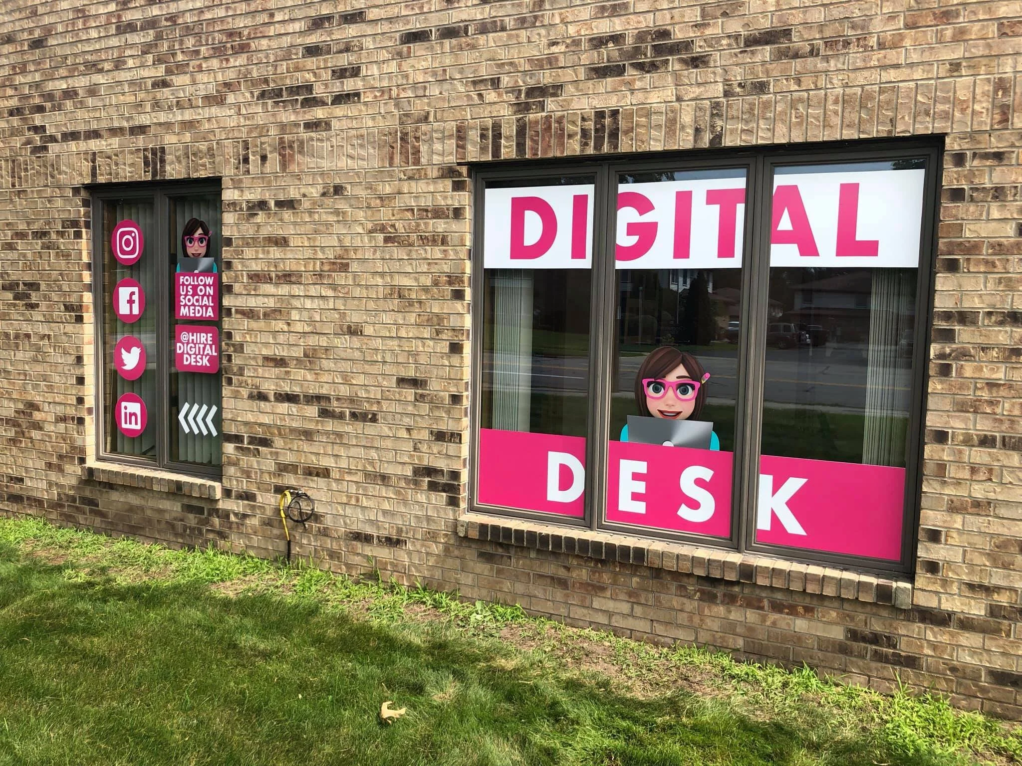Brick building with large windows. One window features digital marketing signs with social media icons and a cartoon character, while the other has a sign that says 'DIGITAL DESK' and a cartoon character with glasses and a laptop.