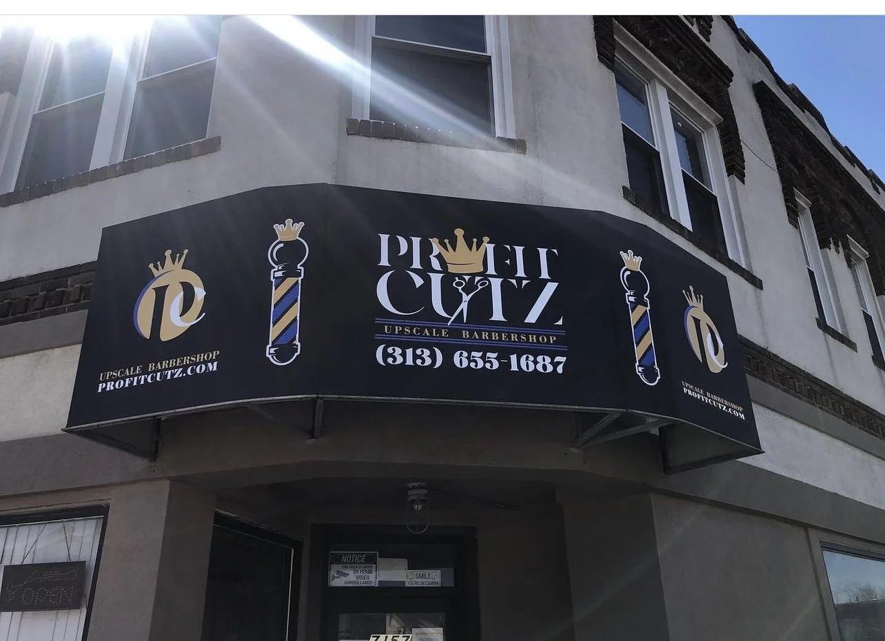 Black awning with gold, white, and blue barber pole graphics and text, promoting a barbershop called Prime Cutz, located on a corner of a building with two floors, multiple windows, and a door.