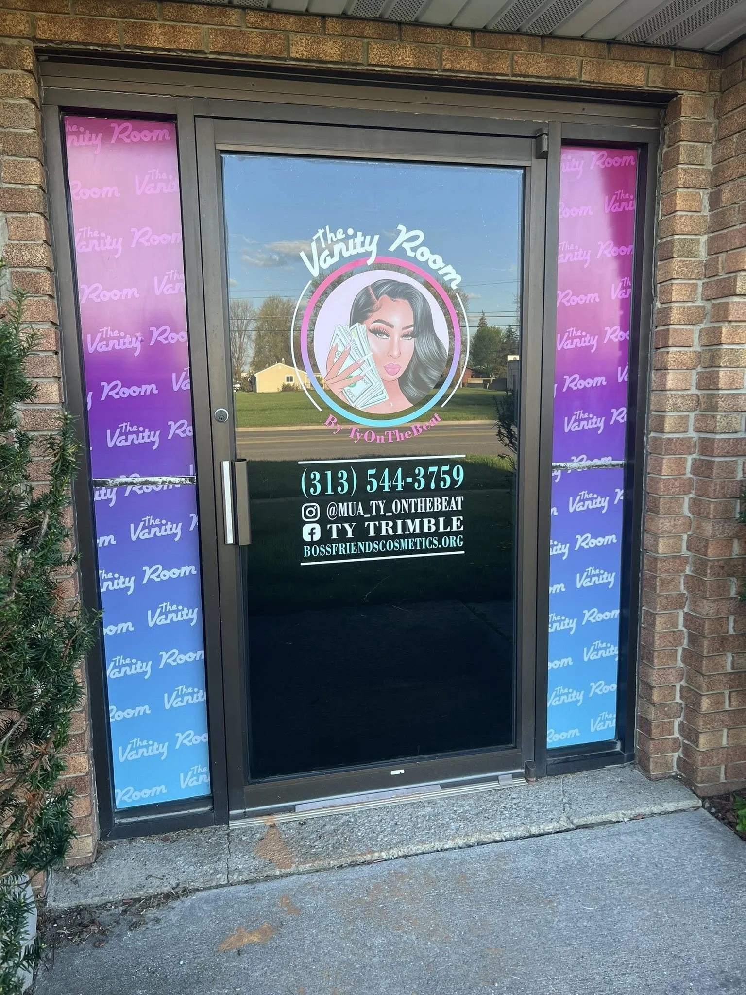 Storefront door with a colorful decal for The Vanity Room, featuring an illustration of a woman with dark hair holding cash, and contact details including phone number, Instagram handle, Facebook page, and website.