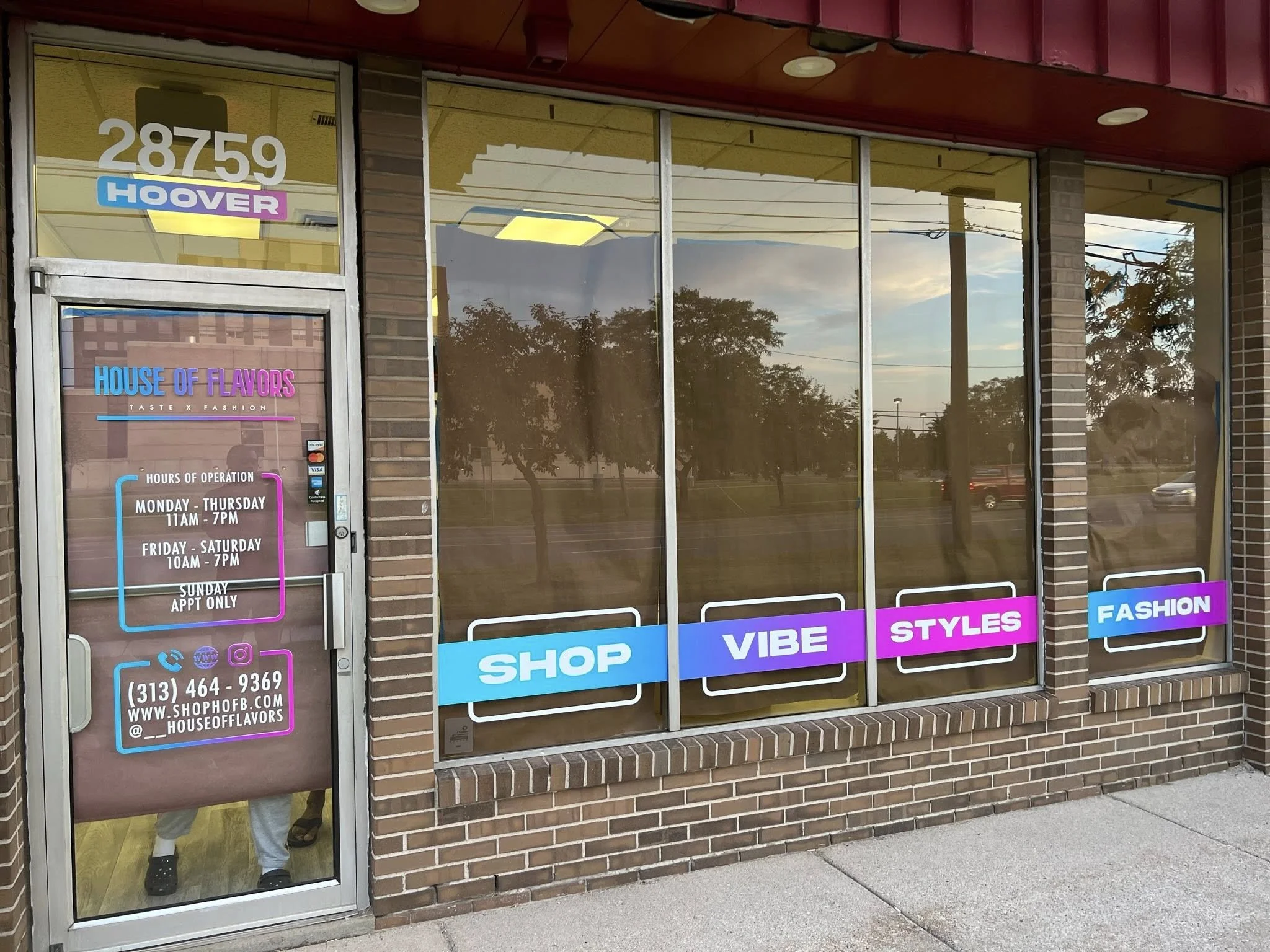 The exterior of a retail store named 'House of Flavors' with large glass windows and a door. The door has signage with business hours, contact information, and social media handles. The store features colorful sign banners on the windows with words: 