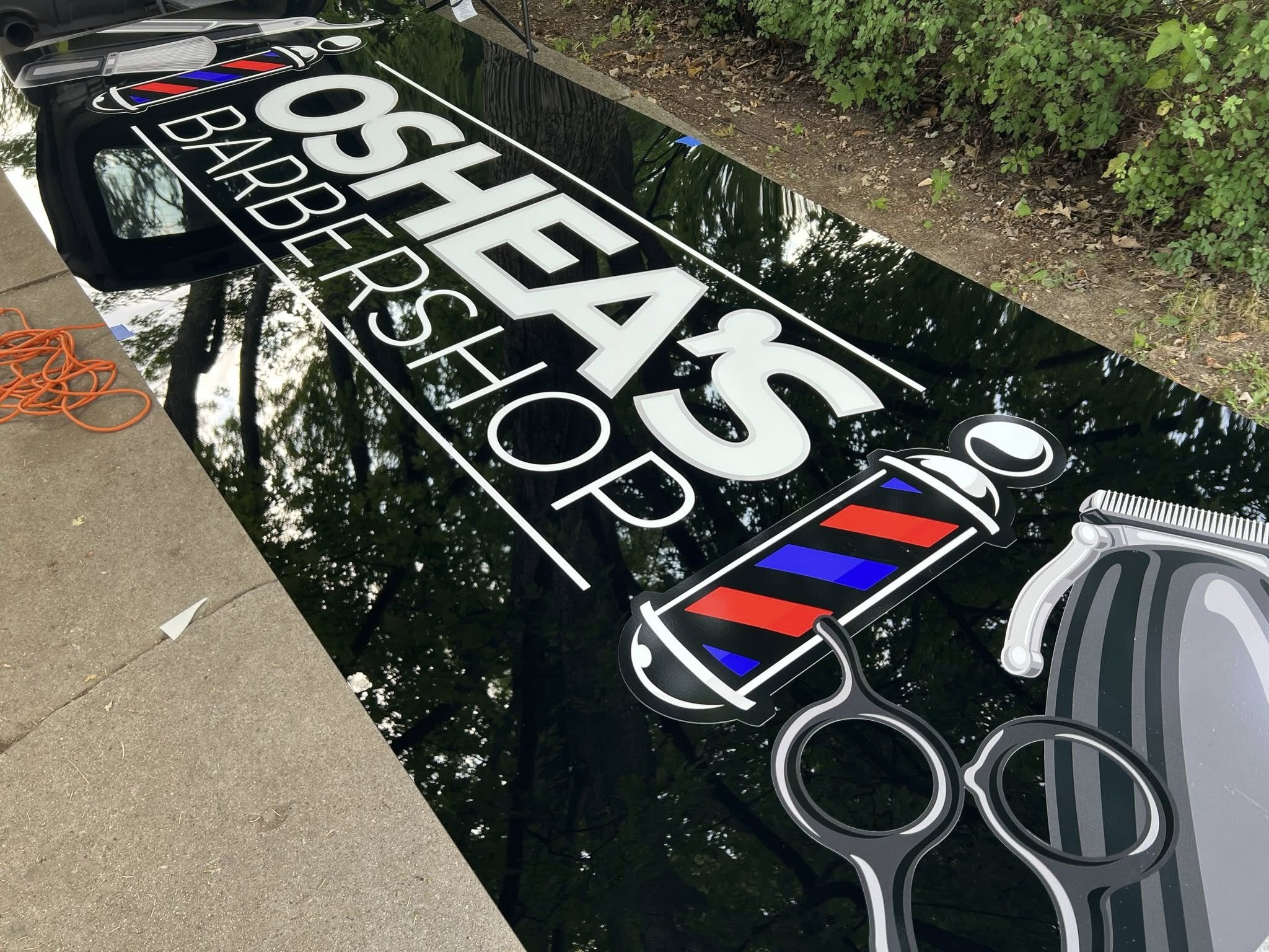 A black sidewalk sign for Osborne Barbershop with cut-out images of a barber pole and hair scissors, placed outdoors on concrete, surrounded by green bushes and trees, with reflections of the sky and trees visible on the glossy surface.