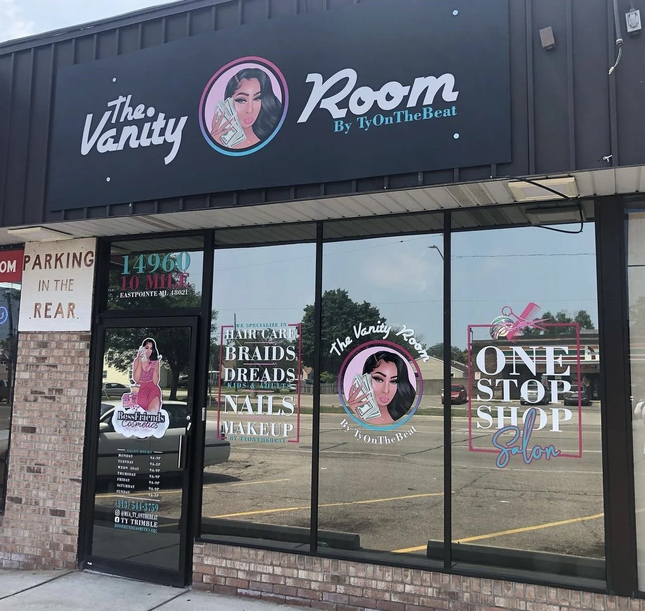 Storefront of The Vanity Room by TyOnTheBeat, a salon offering hair care, braids, dreads, nails, and makeup services. Signage includes a logo with a woman holding money, and text advertising one-stop shop salon. Parking instructions are posted, along