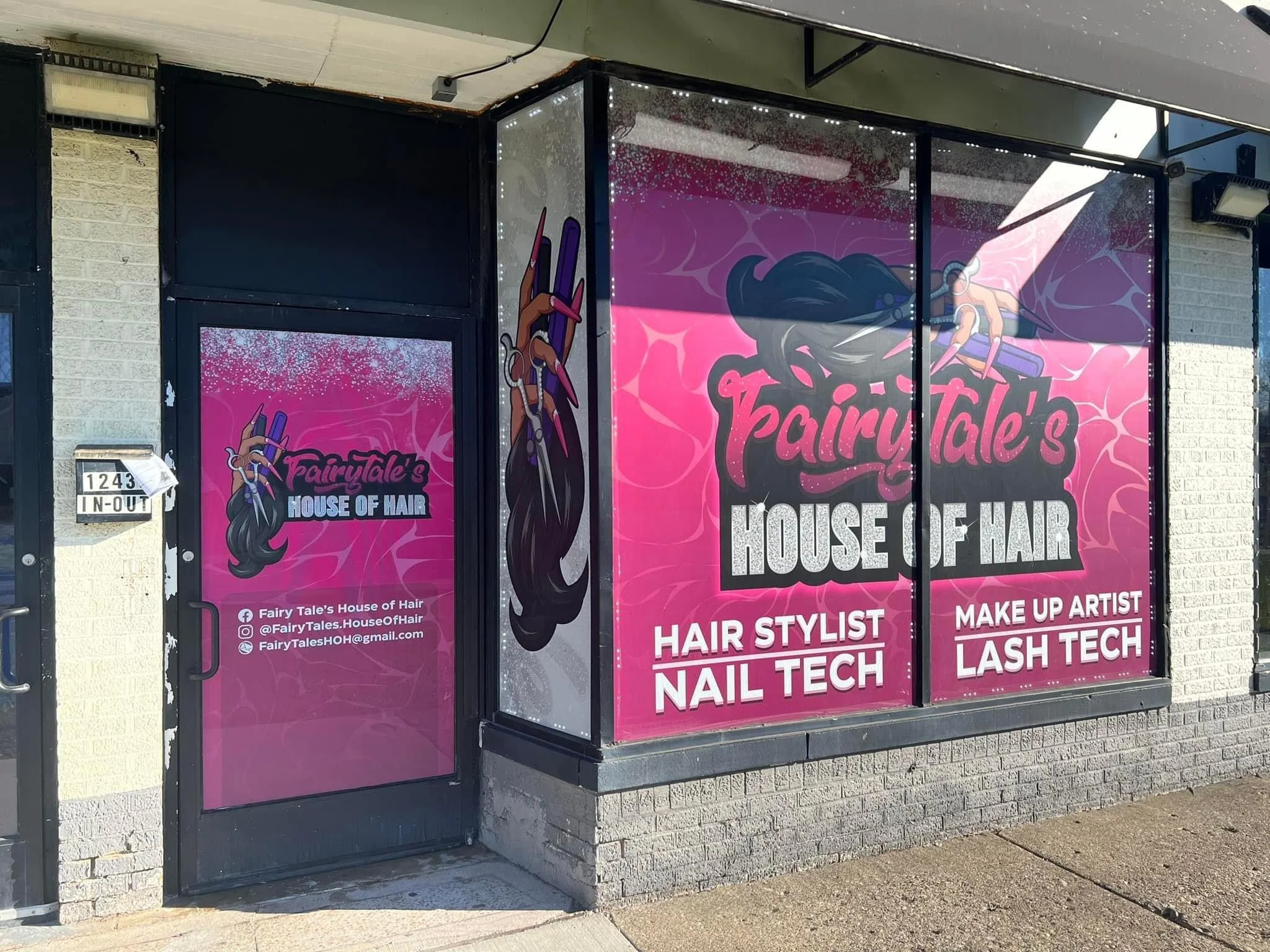 Pink storefront window display for Fairy Tale's House of Hair with a large illustration of a stylized female hand holding a pair of scissors and hair brush. The business offers hair styling, nail tech, makeup artist, lash tech, and information includ