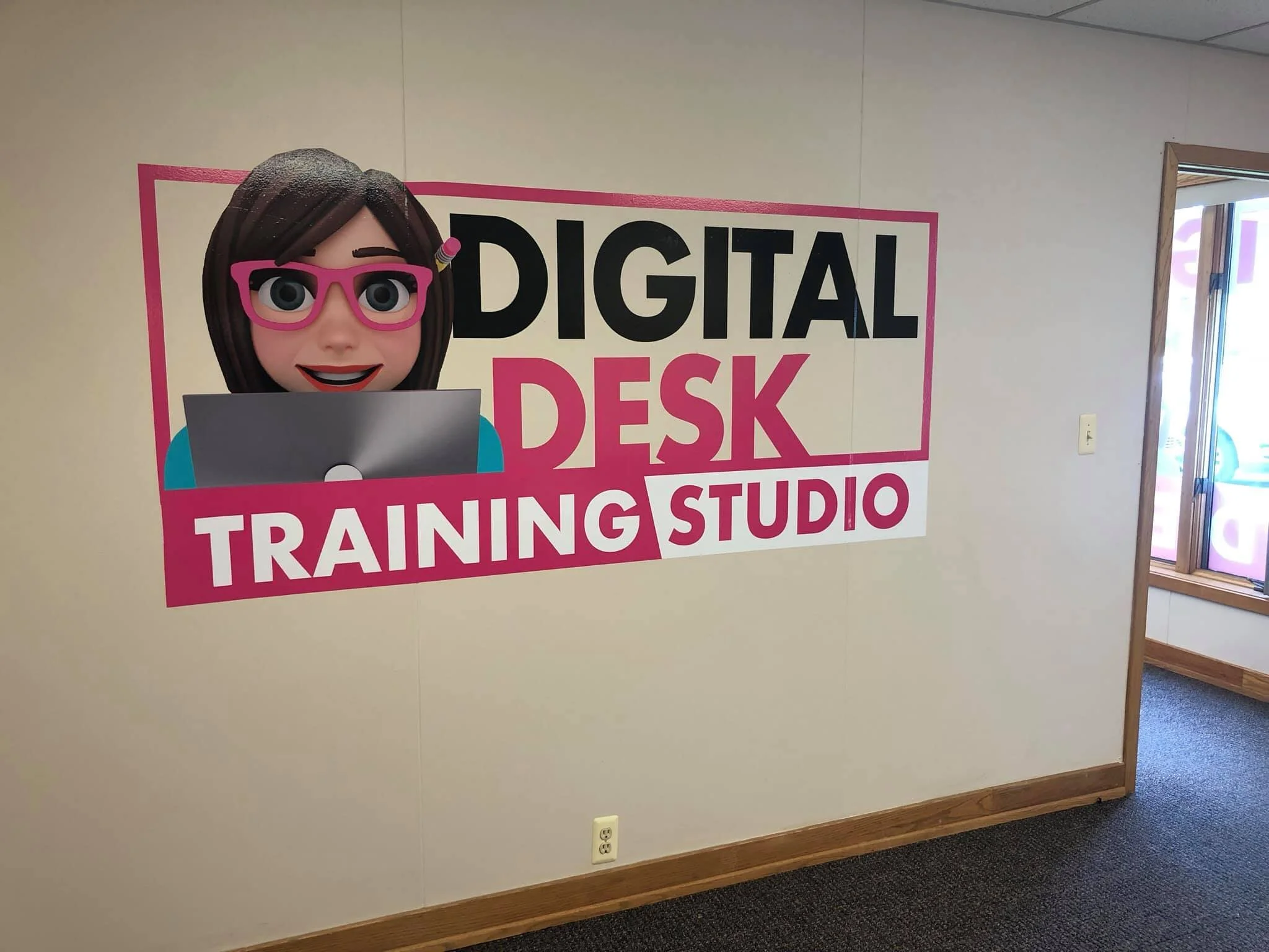 Decorative wall sign reading 'Digital Desk Training Studio' with a cartoon character of a woman with dark hair, pink glasses, and a blue shirt, working on a laptop at a desk.